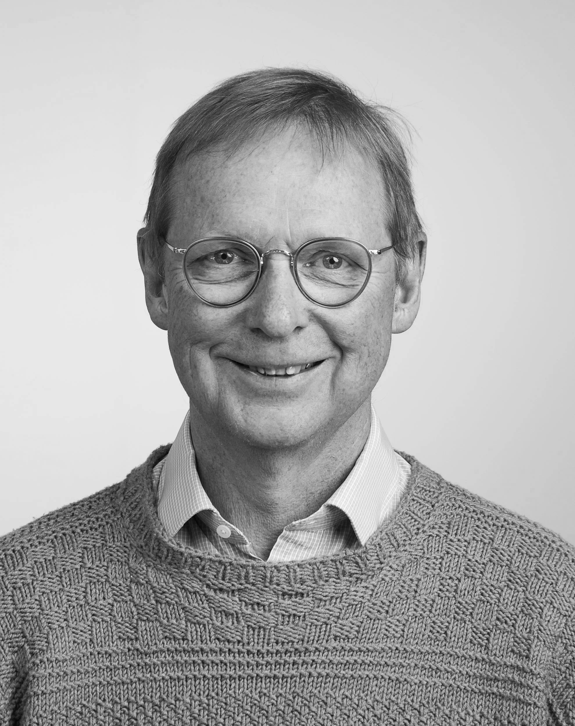 A black and white photo of Gísli Jón Hjaltason, wearing glasses, a sweater and a collared shirt.