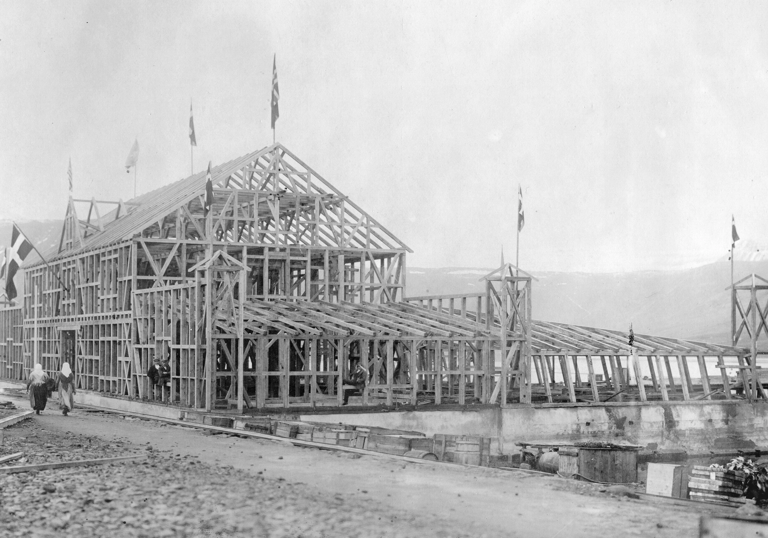 Black and white photo of a large wooden framework of Edinborgarhúsið under construction, with flags atop the structure and several workers visible, set against an open landscape.