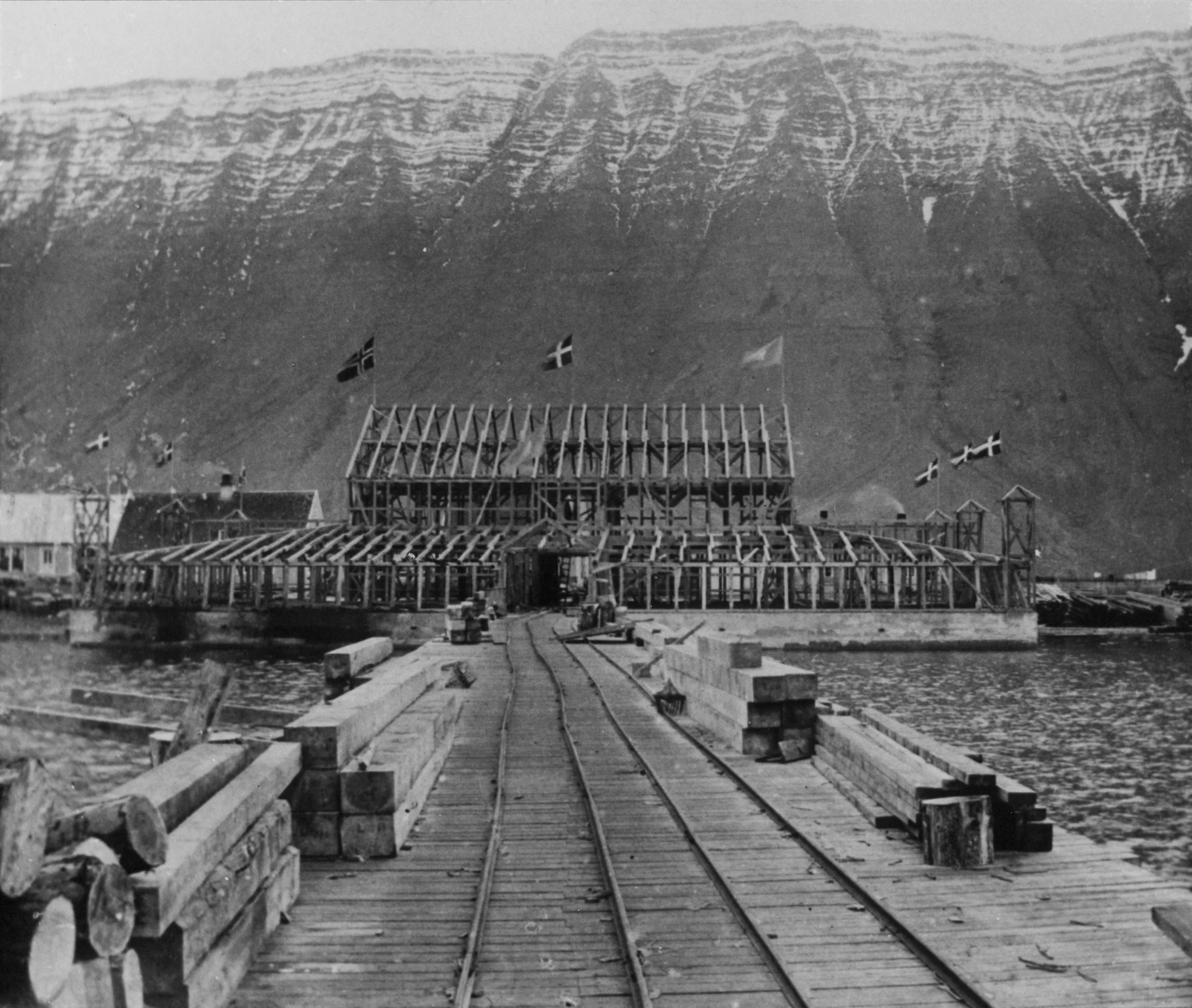 Historical black and white photo of a waterfront construction site with a partially built Edinborgarhúsið, flags flying, and train tracks leading toward the building. The background features a mountain with slight snow cover.