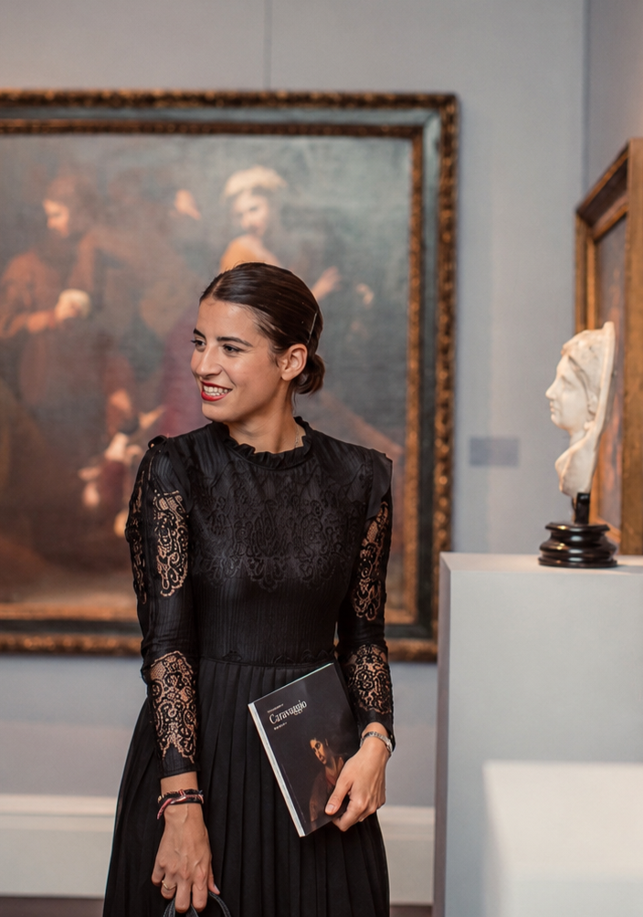 A woman with dark hair tied back, wearing a black lace dress, smiling and holding a booklet, standing in an art gallery with paintings and sculptures in the background.