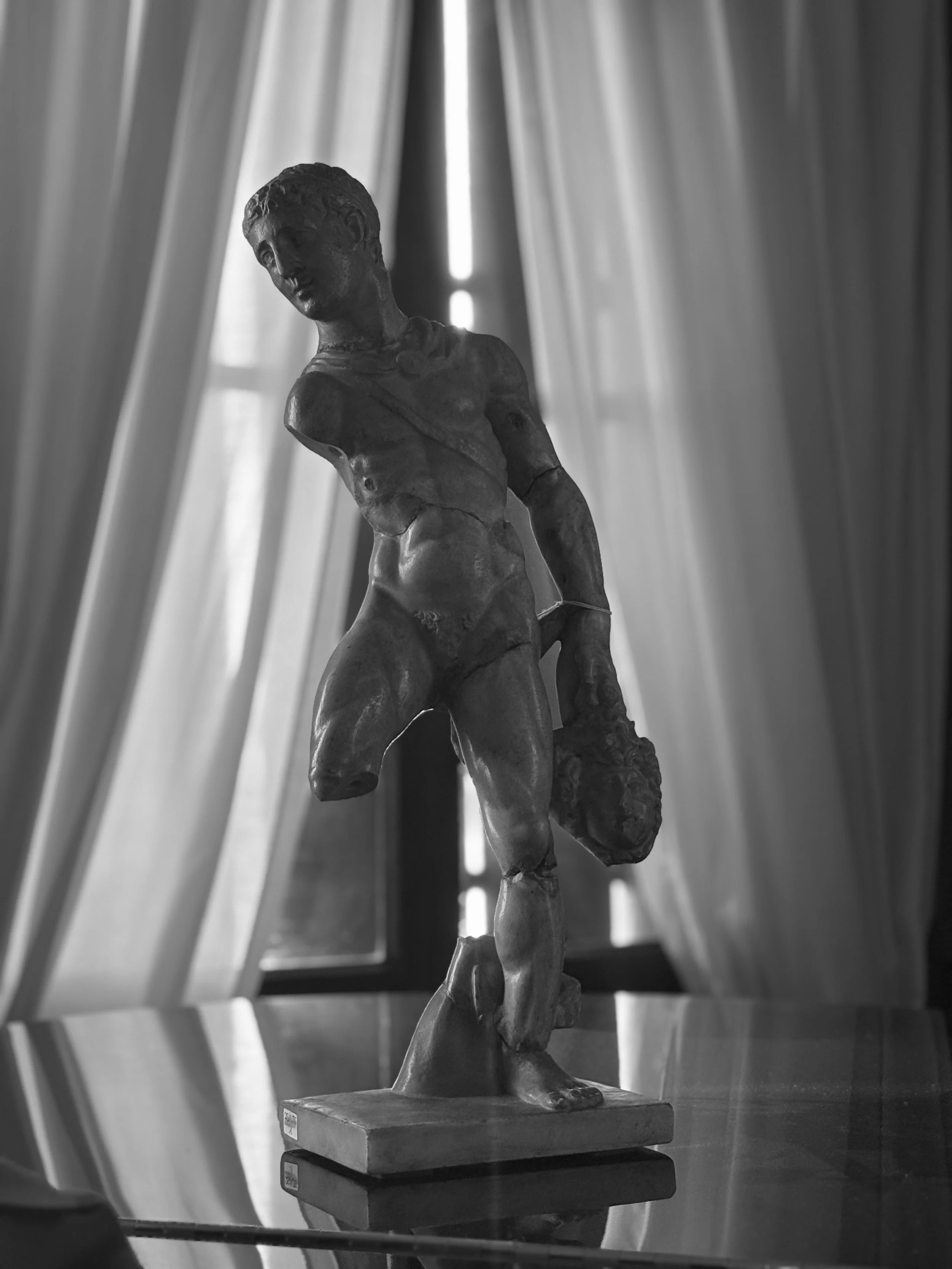 A black-and-white photo of a sculpture of a young man with short hair, bare chest, and an arm band, standing on a base in front of a window with curtains.