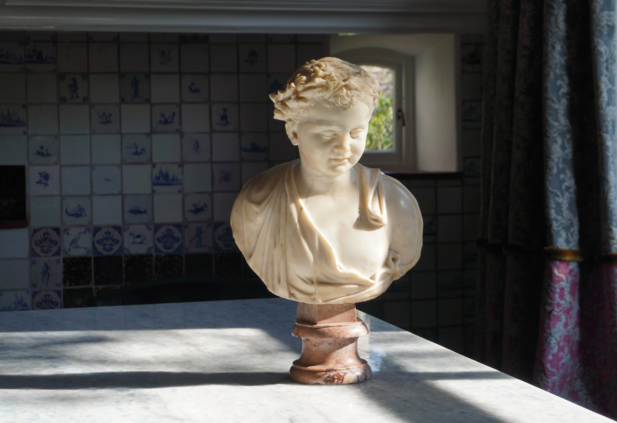 A marble bust sculpture of a young person with curly hair, placed on a marble base, sitting on a marble surface indoors with decorative tile wall and window behind.