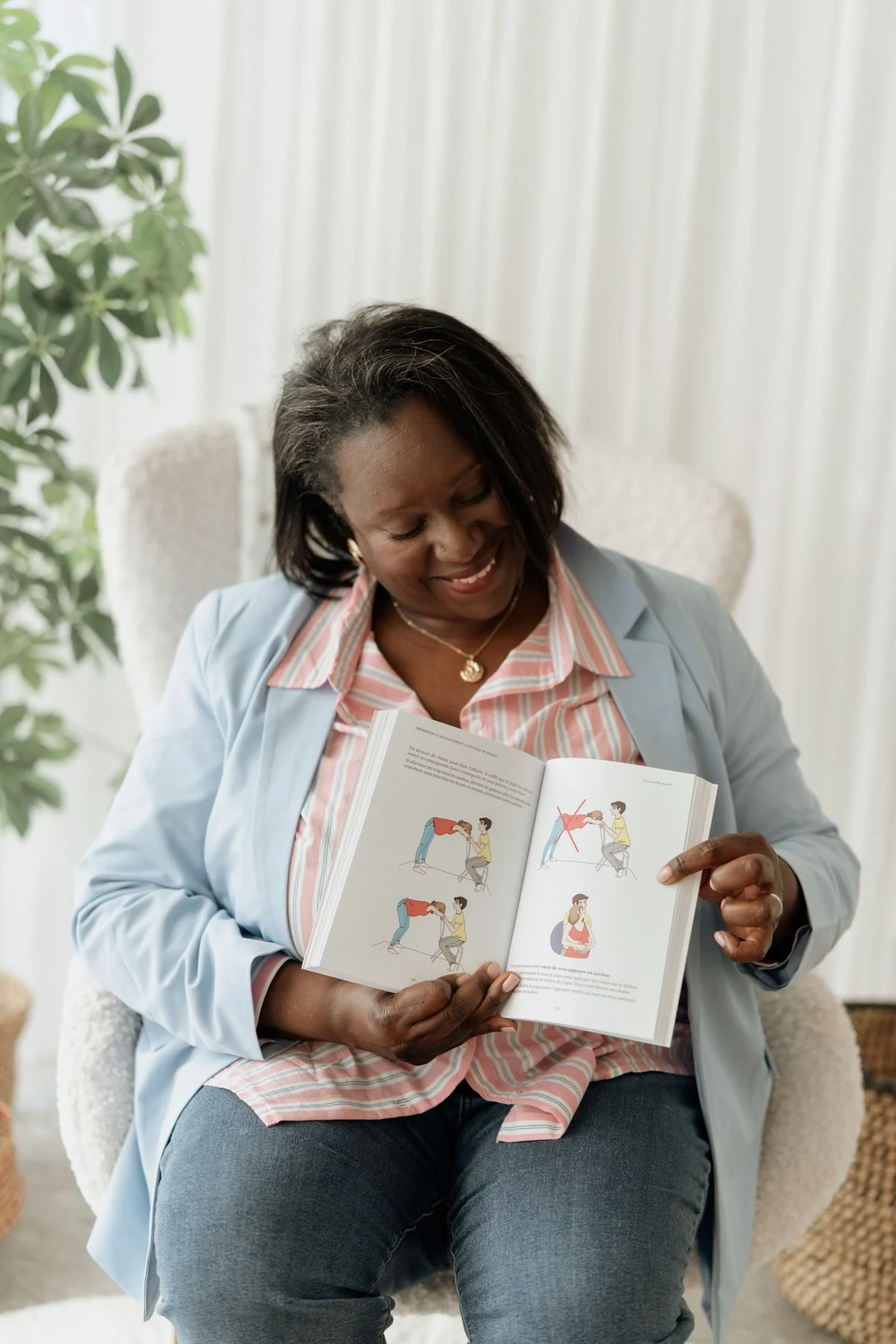 Une femme assise dans un fauteuil, regardant un livre d'instructions sur l'accouchement, avec un fond de rideaux blancs et une plante verte à gauche.