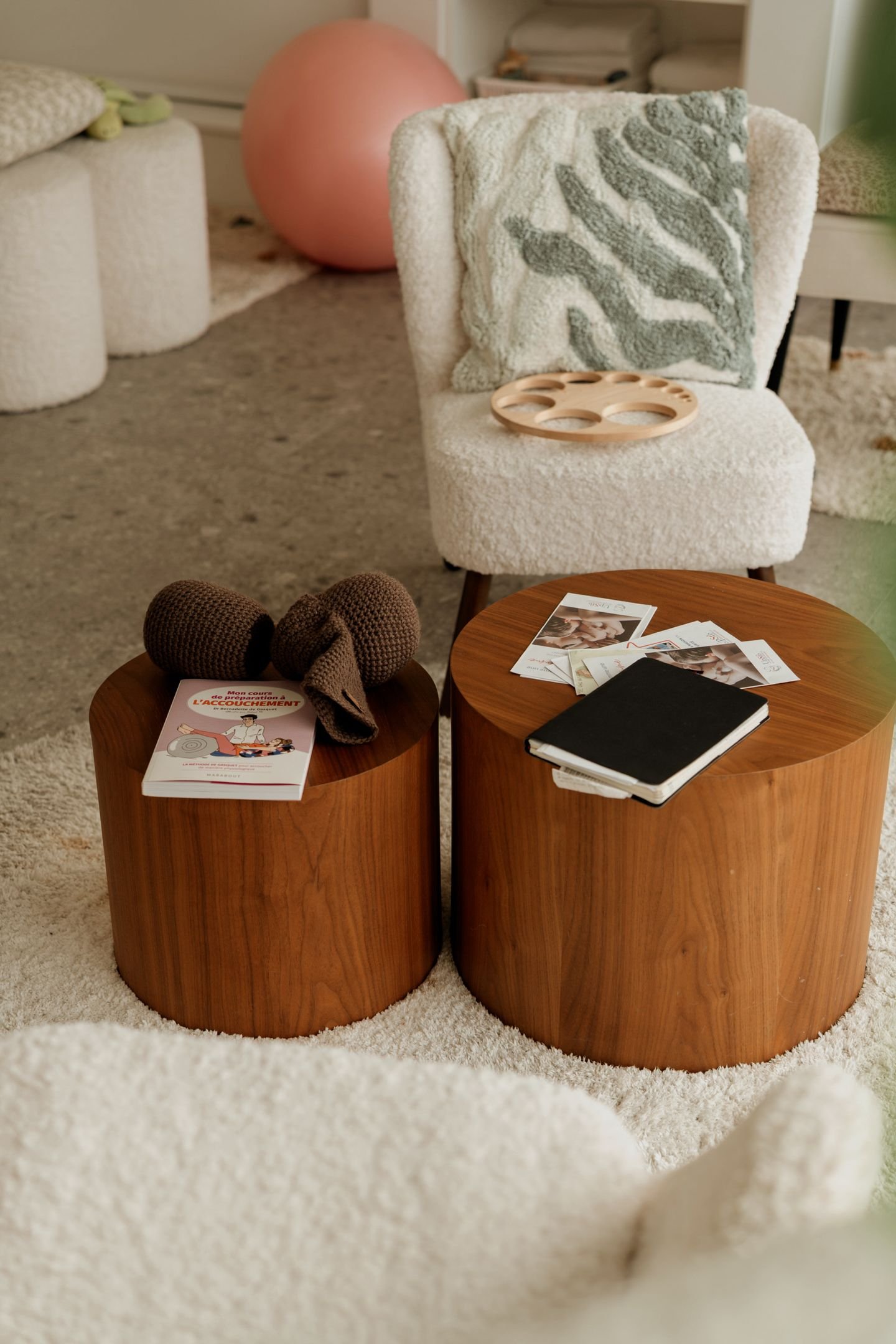 Deux tables en bois rondes avec des livres, un cahier et des pelotes de laine dessus, dans un salon avec deux fauteuils en tissu beige et un coussin à motif de feuilles vertes.