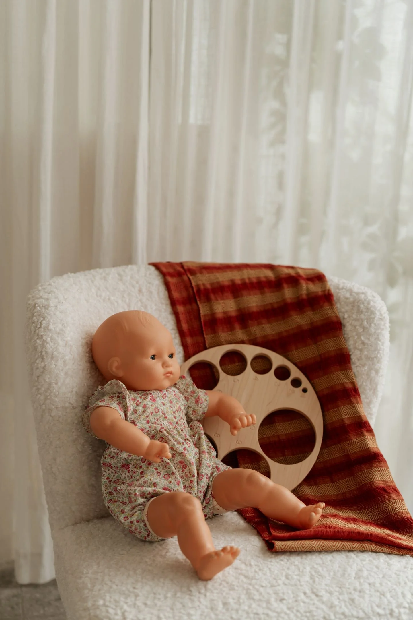 Poupée pour enfants assise sur un fauteuil blanc, avec un disque en bois à côté, derrière un drap blanc et une couverture à carreaux rouges et marrons.