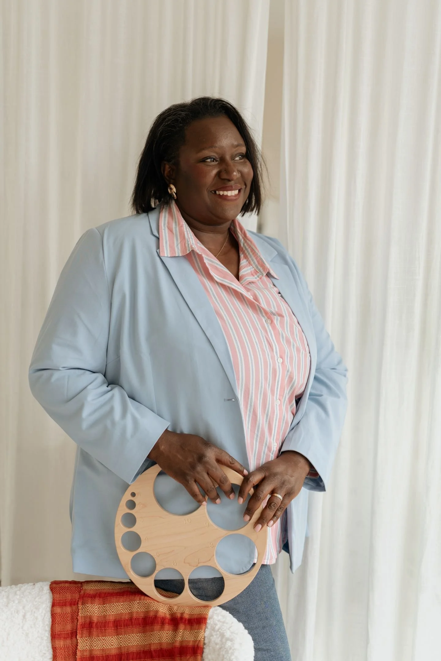 Une femme souriante qui tient un objet en forme de disque avec des trous, portant une veste bleue et une chemise à rayures rose et blanche, avec un rideau blanc en arrière-plan.