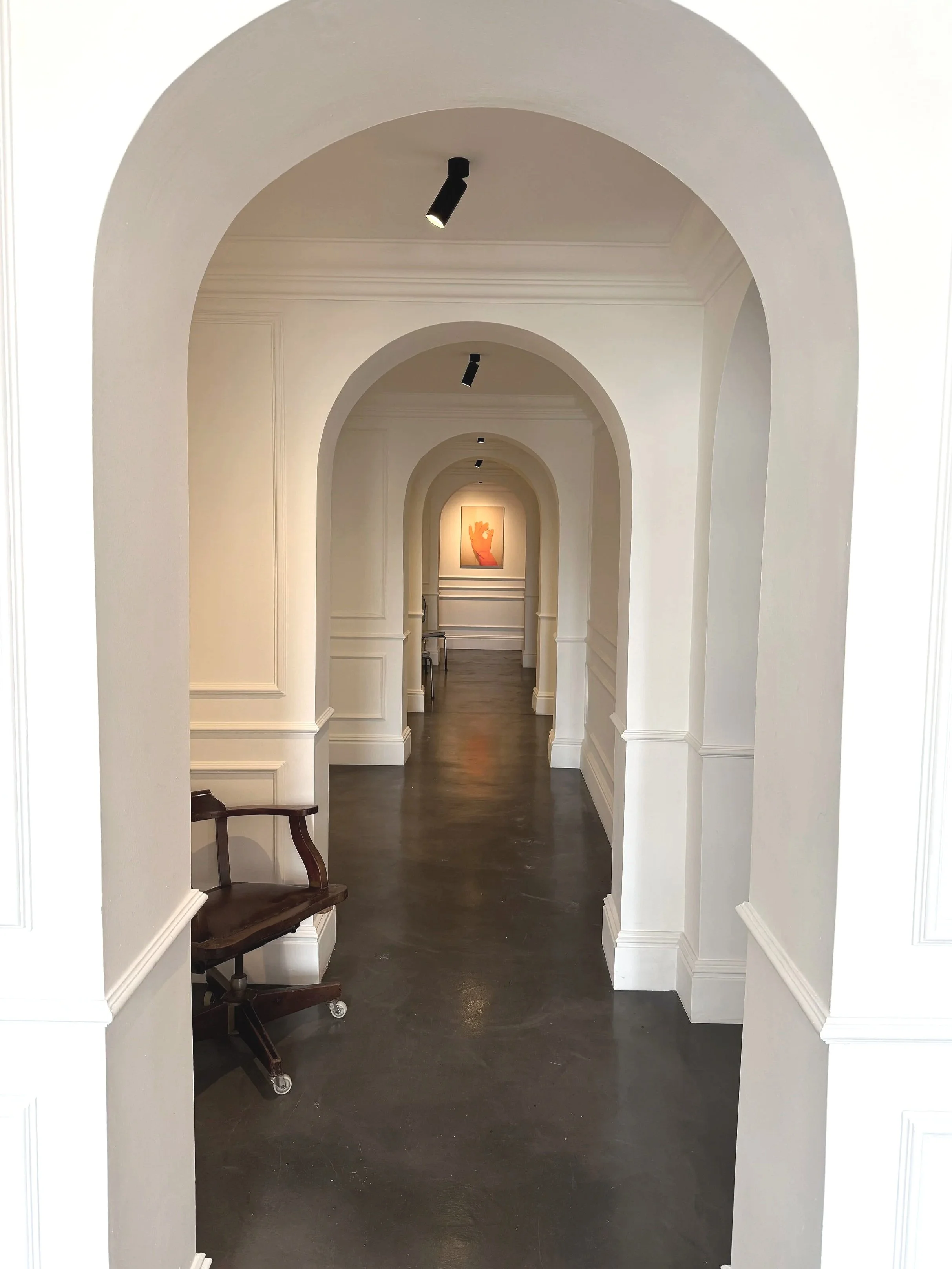 A hallway with multiple white arches and black ceiling lights, with a dark polished floor and a framed artwork at the end depicting an orange hand gesture.