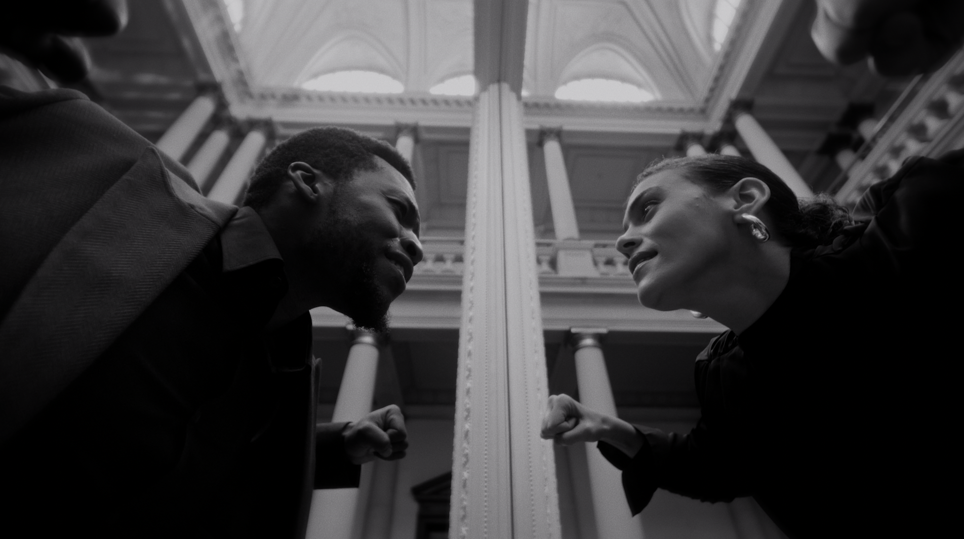 A black and white photo of a man and woman facing each other, both with clenched fists, showing confrontation or argument in an indoor setting with tall columns and arched ceilings.