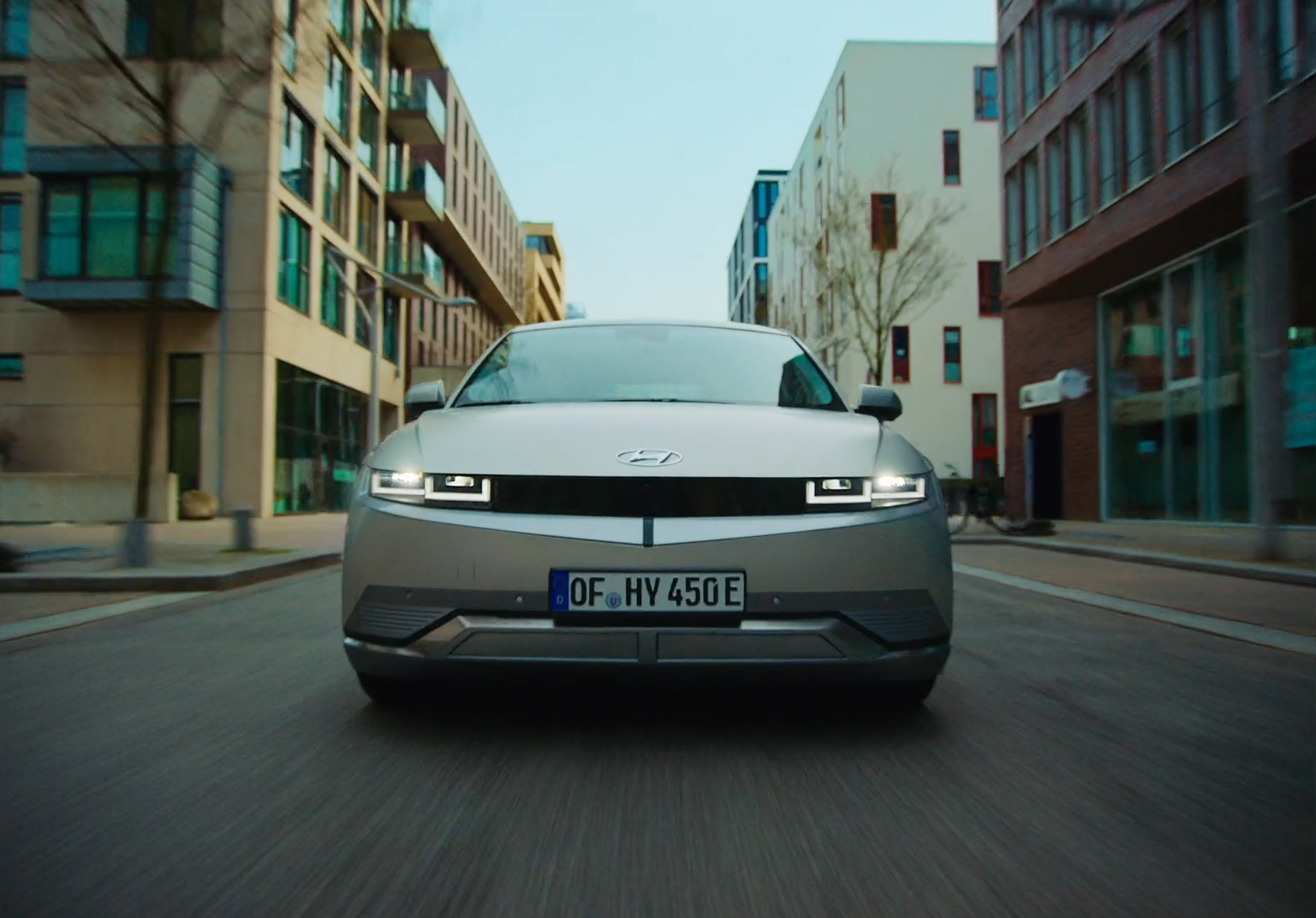 Front view of a silver modern electric car driving through a city street with buildings on both sides.