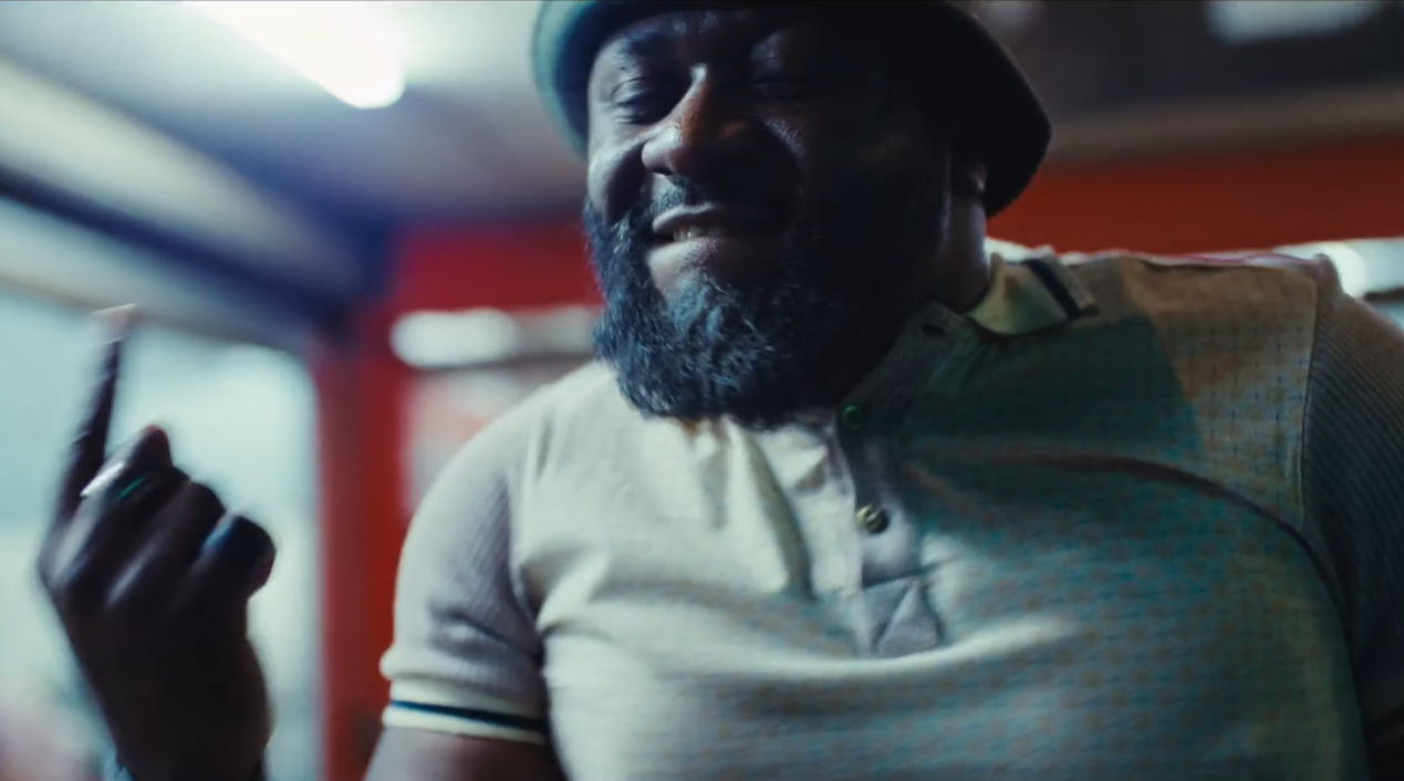 Man with a beard and a hat, smiling and raising his finger in a lively setting.