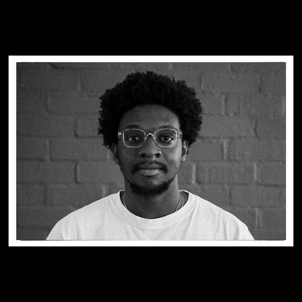 Black and white portrait of a young man with glasses, curly hair, and a faint beard, wearing a white t-shirt, against a brick wall background.