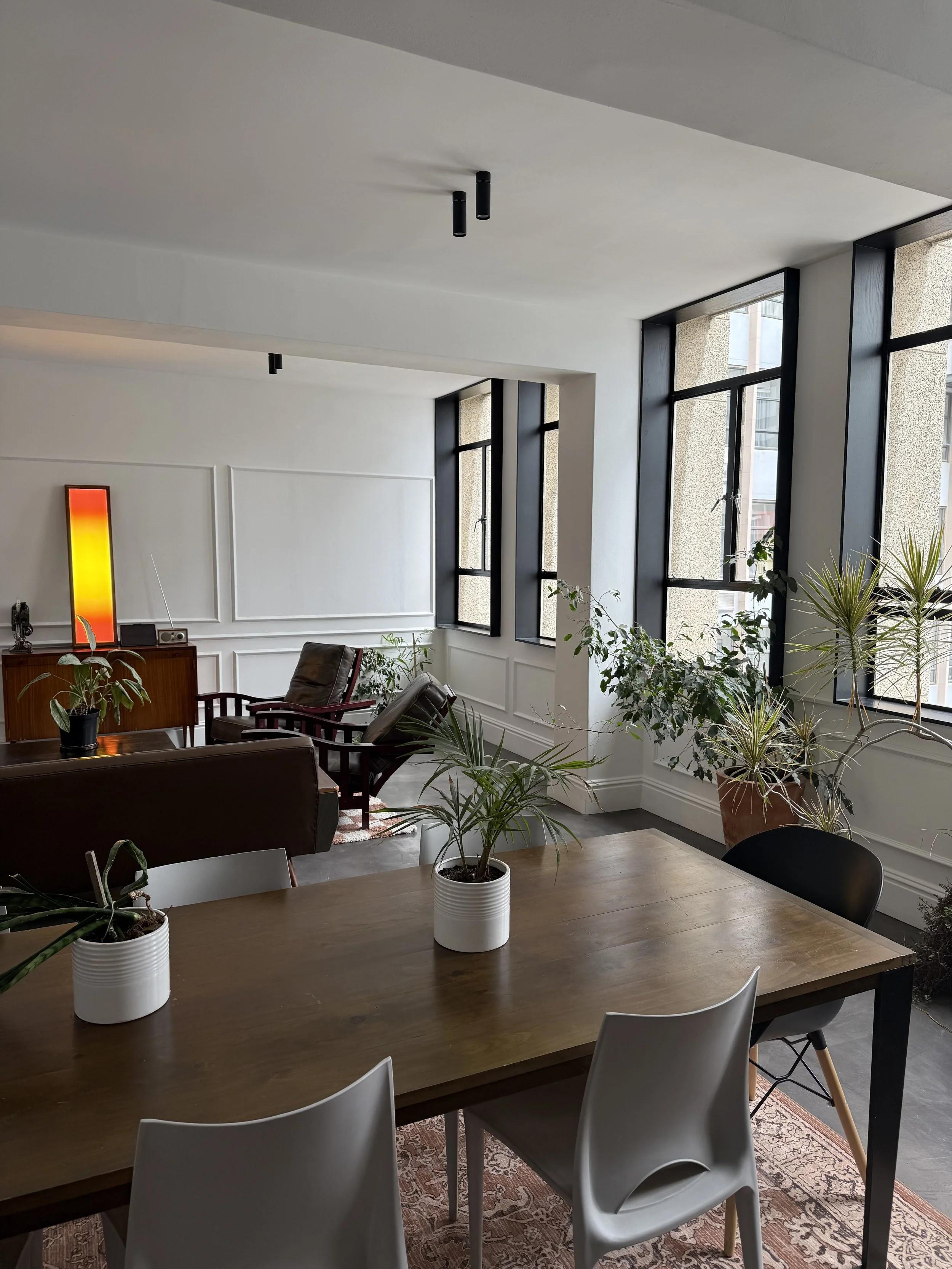 Modern interior living space with large windows, white walls, a wooden dining table with potted plants, and a sitting area with armchairs and a sideboard.