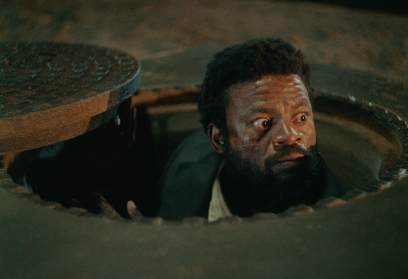 A man with a beard and short curly hair looking upward from a manhole, with a surprised or concerned expression, in a scene with a dark, gritty background.