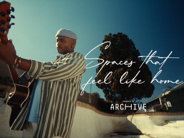 A young man with a beanie and striped shirt playing an acoustic guitar outdoors under a large tree on a sunny day, with text overlay that reads 'Spaces that feel like home' and 'Presented by ARCHIVE'.