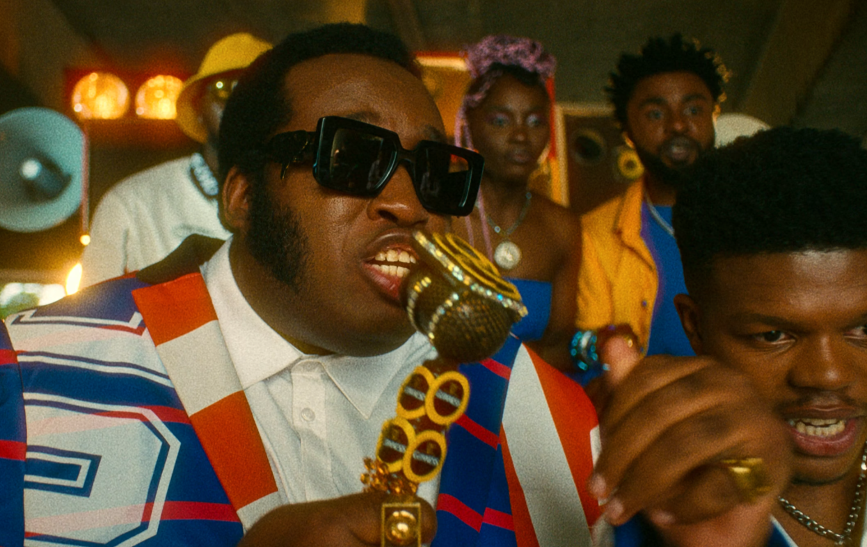 A man in sunglasses singing into a gold microphone in a colorful party setting, wearing a patterned jacket.