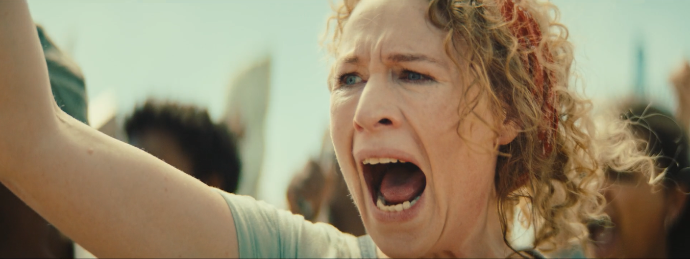 A woman with curly red hair shouting with her mouth open and eyes wide, surrounded by a crowd.