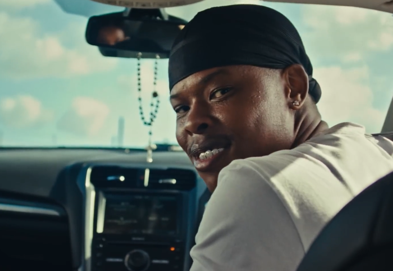 A man with a black durag sitting in the driver's seat of a vehicle, looking back and smiling.