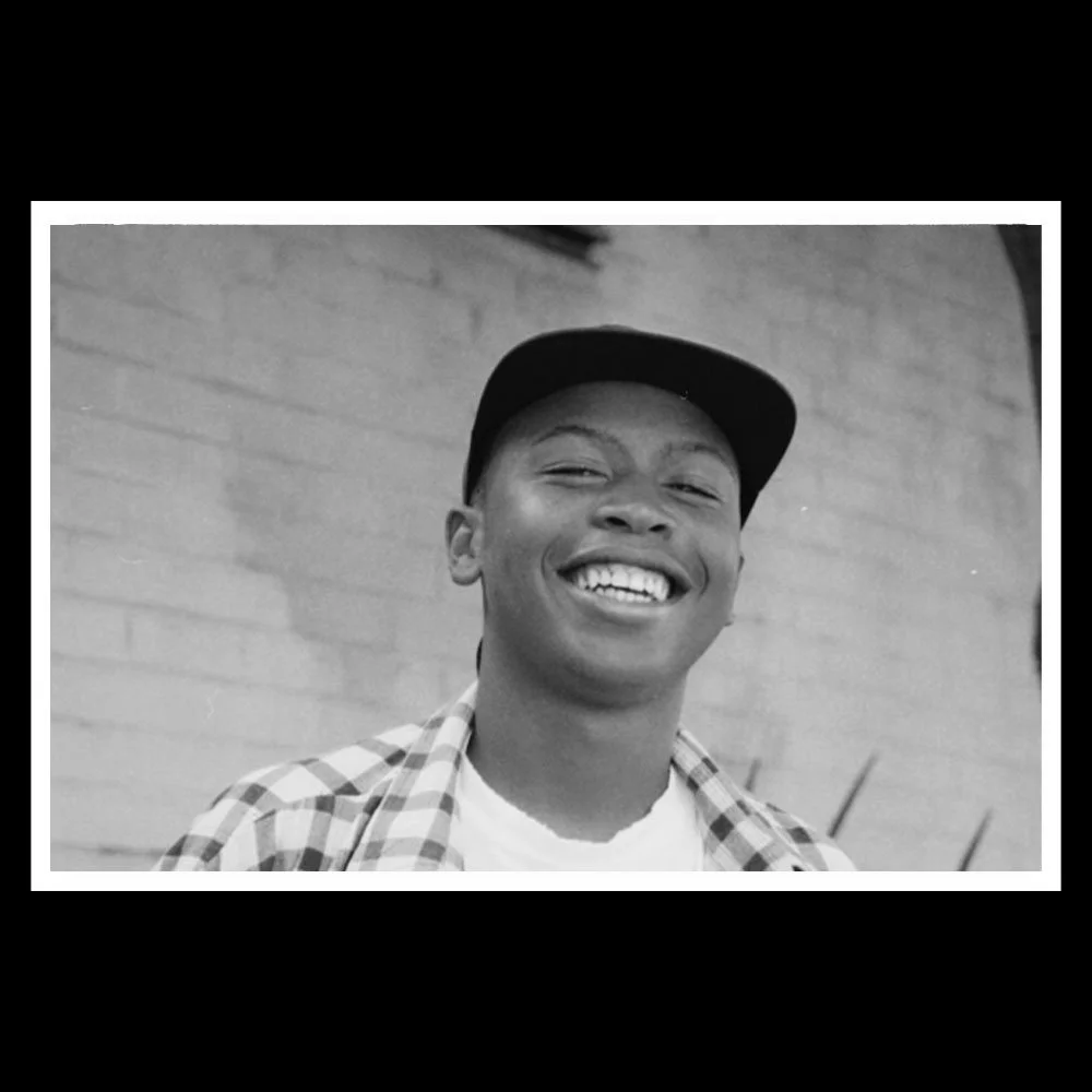 Black and white photo of a smiling young man wearing a baseball cap and a checkered shirt, standing outdoors with a brick wall in the background.