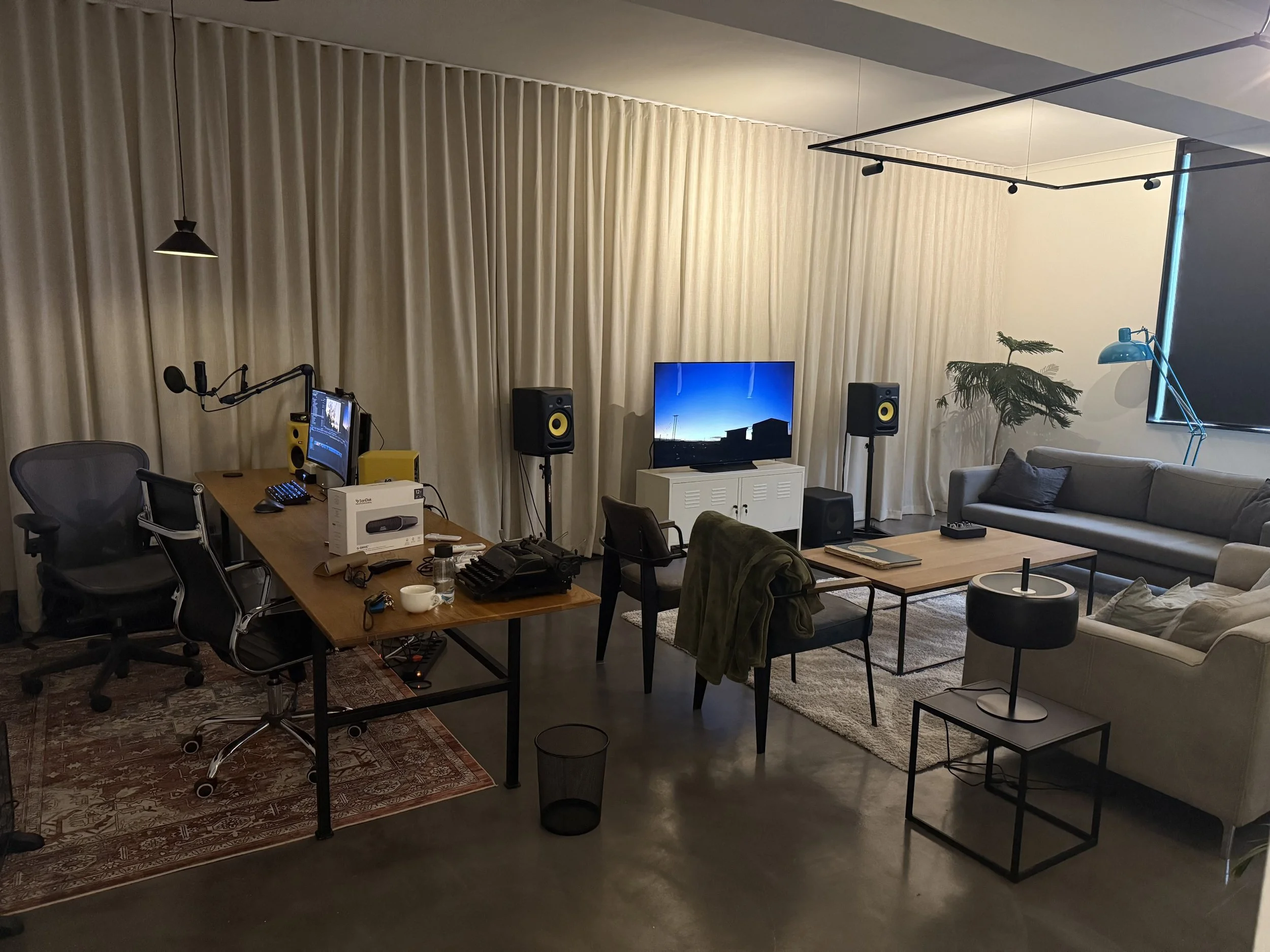 A modern, cozy home office and entertainment area with a large beige couch, a TV on a white stand, a wooden coffee table, a desk with computer equipment, a microphone, and sound speakers, all against a backdrop of beige curtains and a ceiling light, with a large window on the right and a blue desk lamp.
