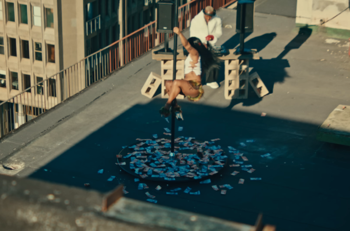 A woman in a revealing outfit dancing on a pole on a rooftop. Someone is sitting nearby in front of a staircase.
