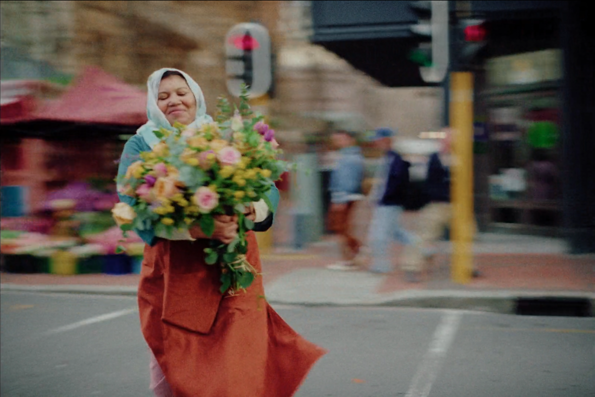 A woman wearing a headscarf holding a large bouquet of colorful flowers walking on a city street with blurred pedestrians in the background.