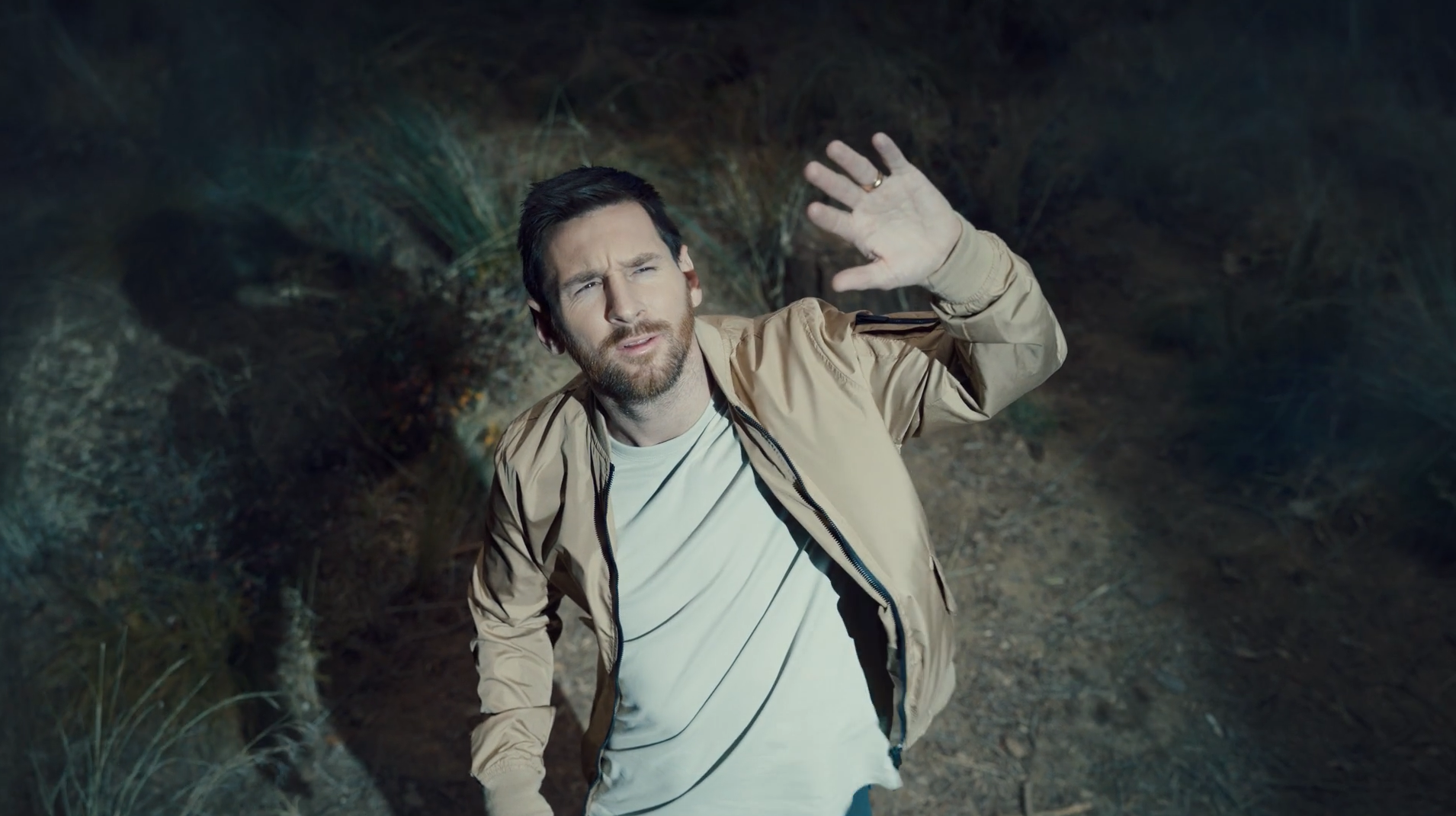 A man with dark hair and a beard is outdoors at night, raising his hand in a wave or to shield his eyes, with a questioning or confused expression on his face.