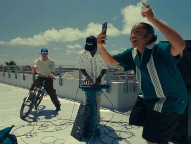 Three young people enjoying music and dancing outdoors on a rooftop on a sunny day, with one person playing a keyboard and another taking a selfie.