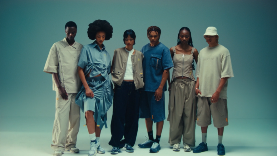 Group of six young people standing together in a minimalist studio setting, dressed in casual, streetwear-style clothing with neutral and pastel colors.