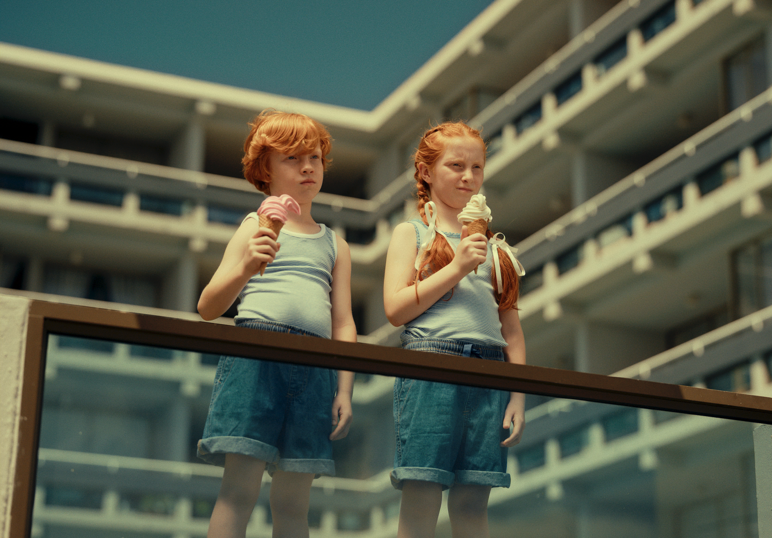 Two young girls with red hair wearing striped tank tops and denim shorts hold ice cream cones, standing outside with a large multi-story hotel or apartment building in the background.
