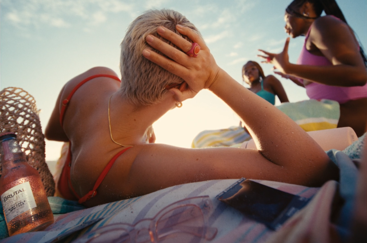 A woman with short blonde hair and a red swimsuit lying on a beach towel, shielding her eyes from the sun with her hand. Two women in swimsuits are standing and talking nearby. Beach items, including a bottle, sunglasses, and magazines, are scattered around.