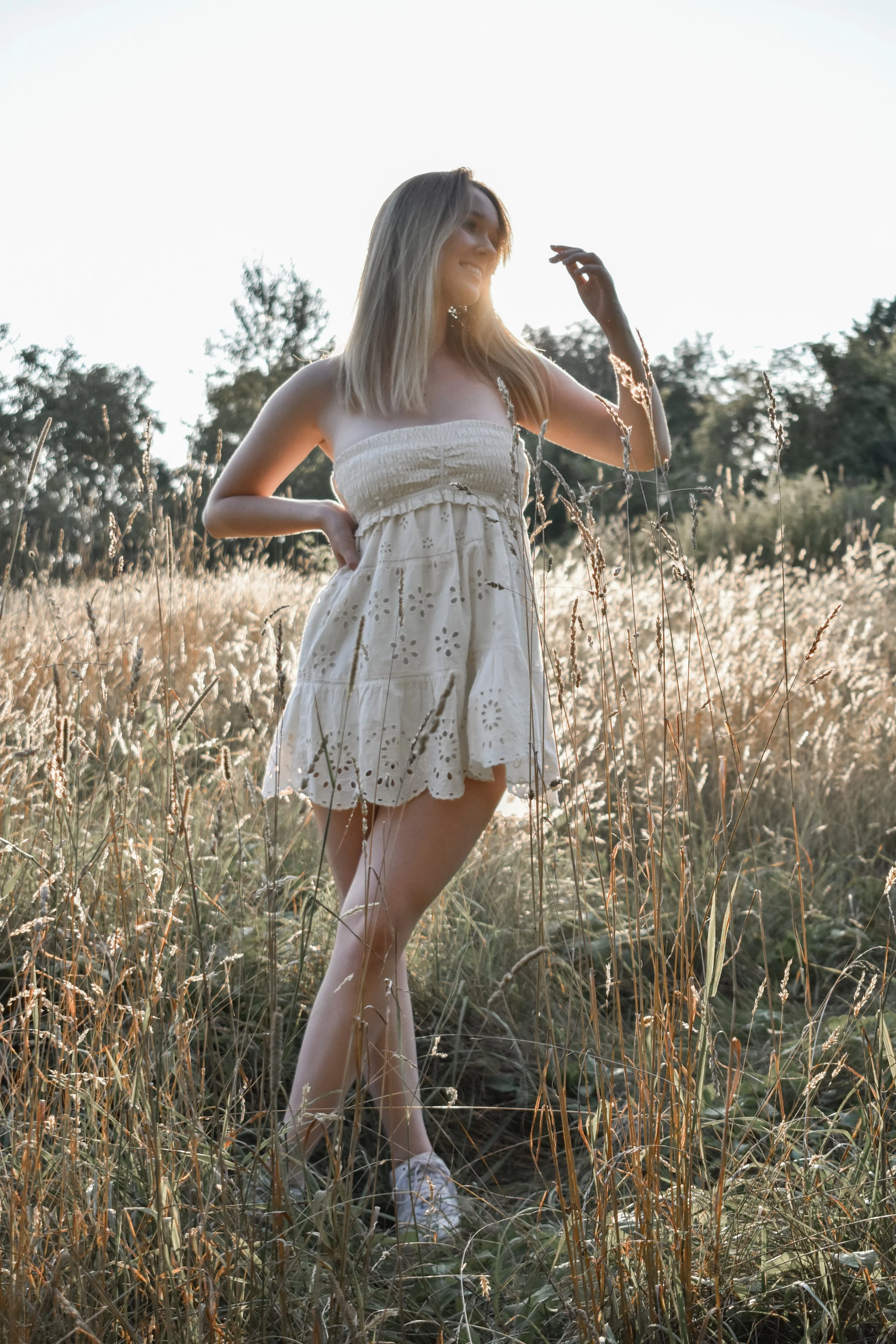 A young woman in a white dress standing in a field of tall grass during sunset.
