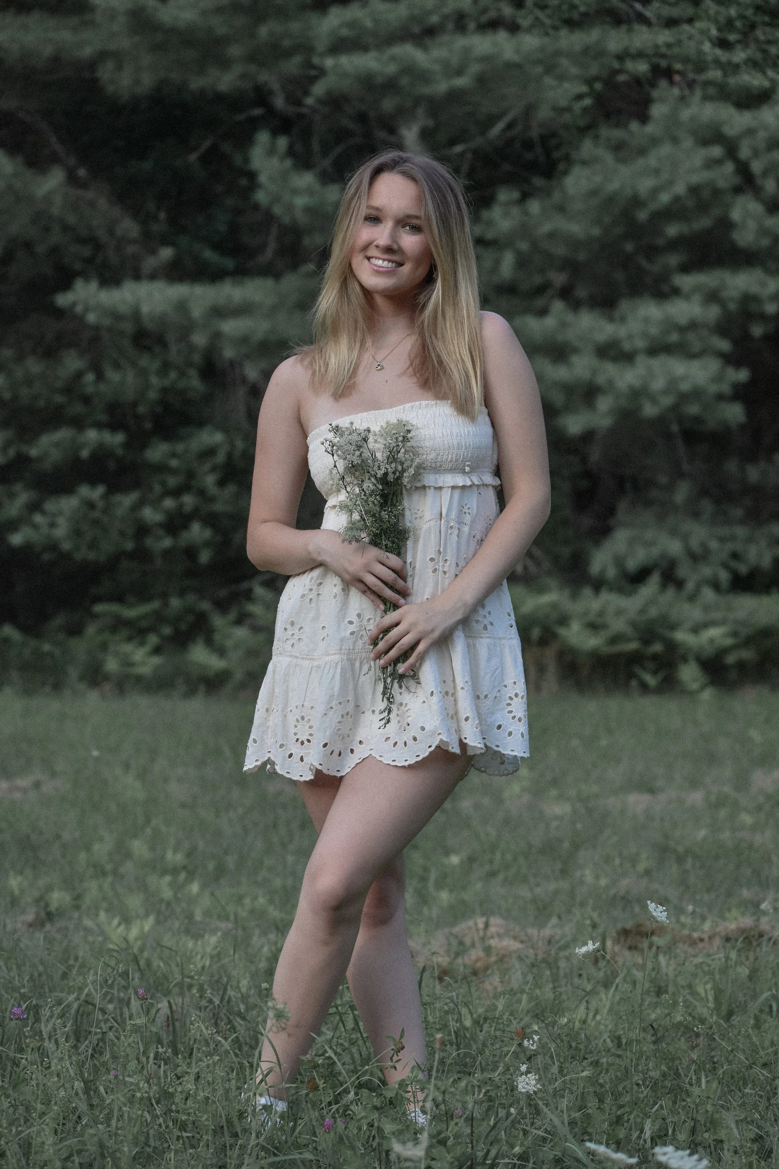 A young woman with blonde hair smiling and holding a small bouquet of wildflowers, standing outdoors on grass with trees in the background, wearing a white sleeveless dress.