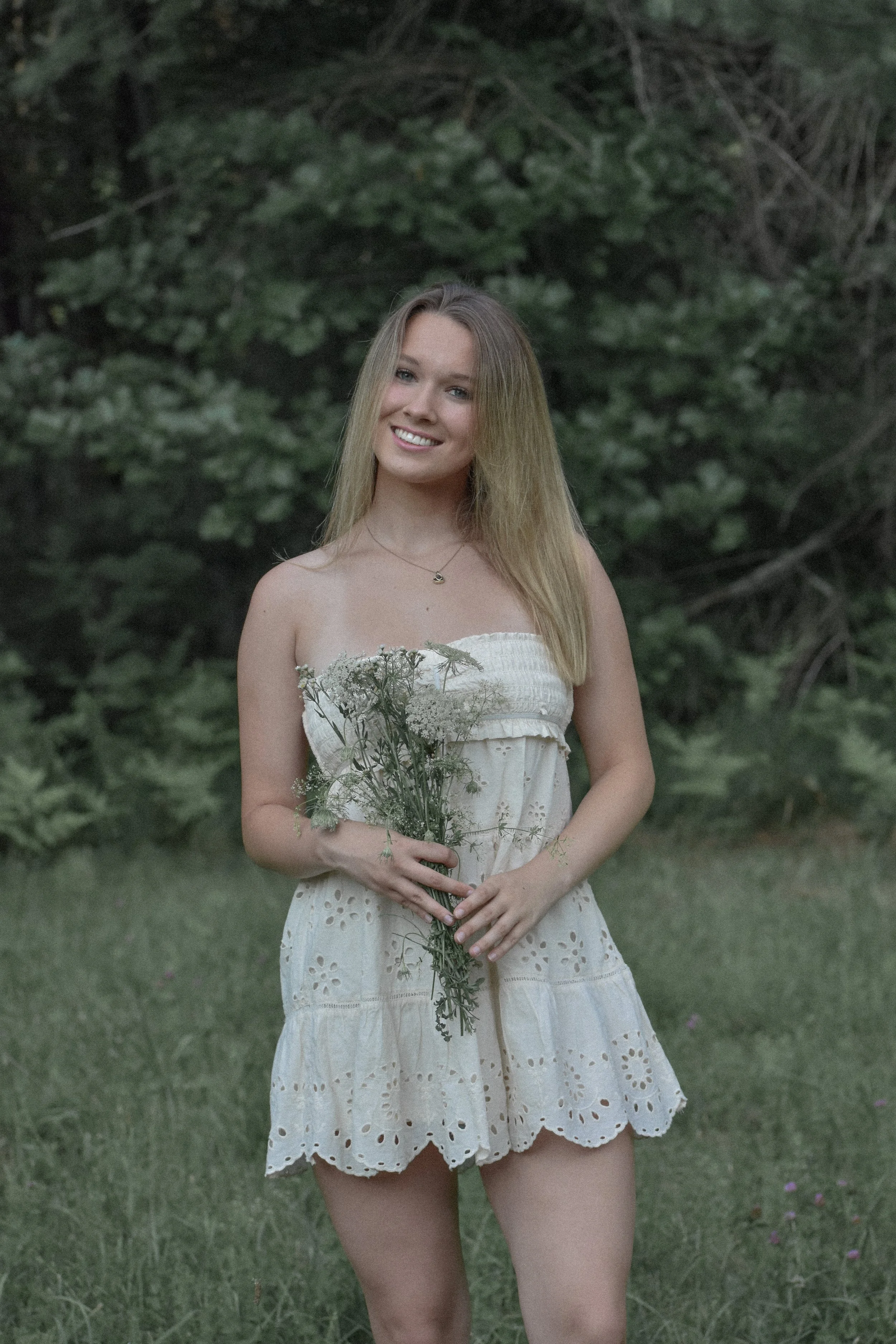 A young woman in a white eyelet dress holding a bouquet of wildflowers stands outdoors in front of greenery, smiling at the camera.