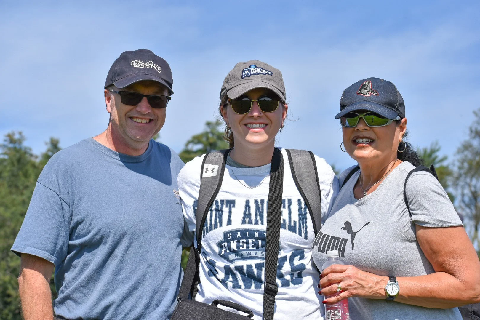 Three smiling people standing outdoors under a blue sky, wearing casual clothes and sunglasses, with trees in the background.