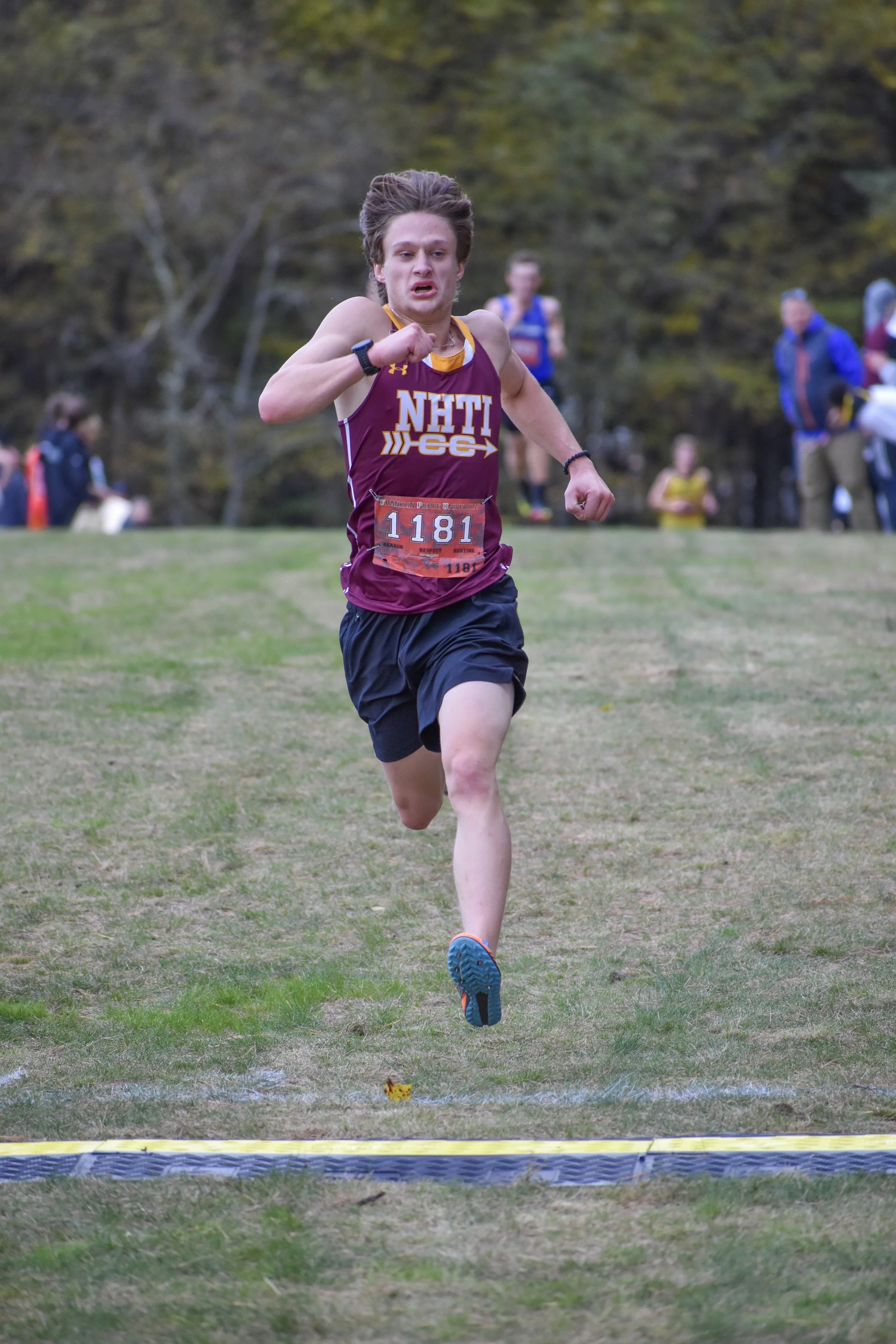 A male runner in a maroon and yellow athletic uniform with 'NHTI' and 'GEC' written on it, racing across a grassy field during a cross-country meet. He is wearing a race bib number 1181 and is approaching the finish line, with others and trees in the