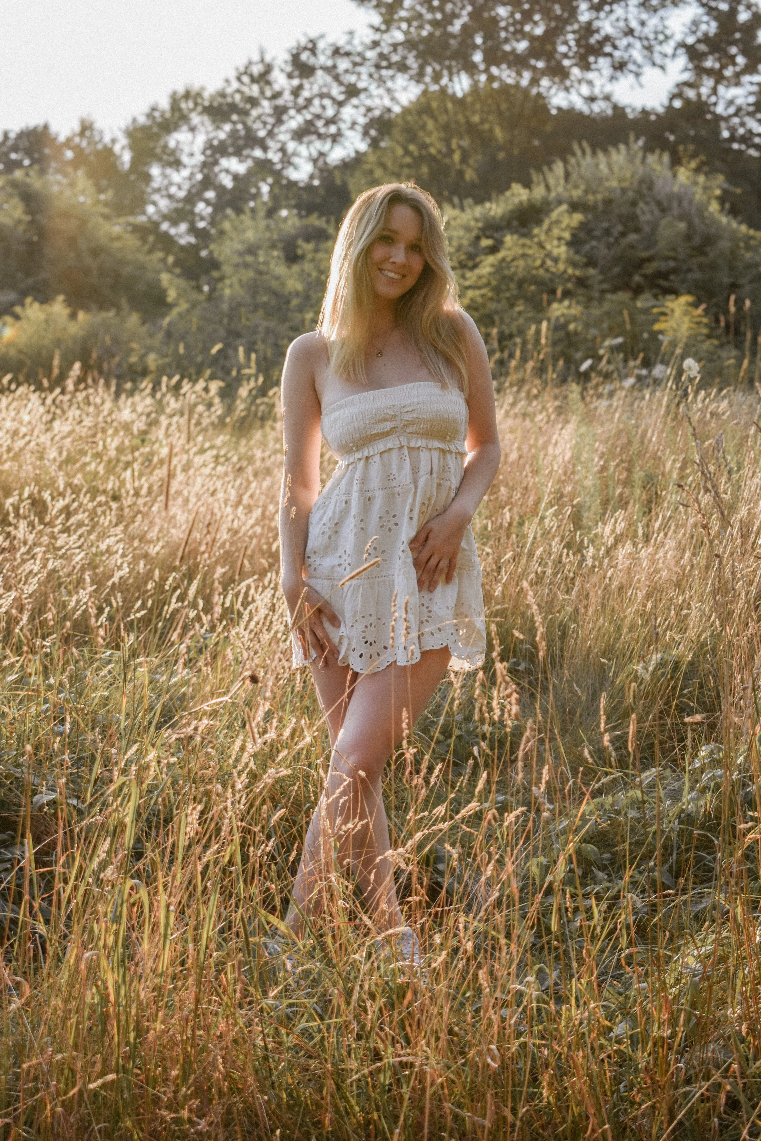 A young woman in a white sundress standing in a field of tall grass during sunset.