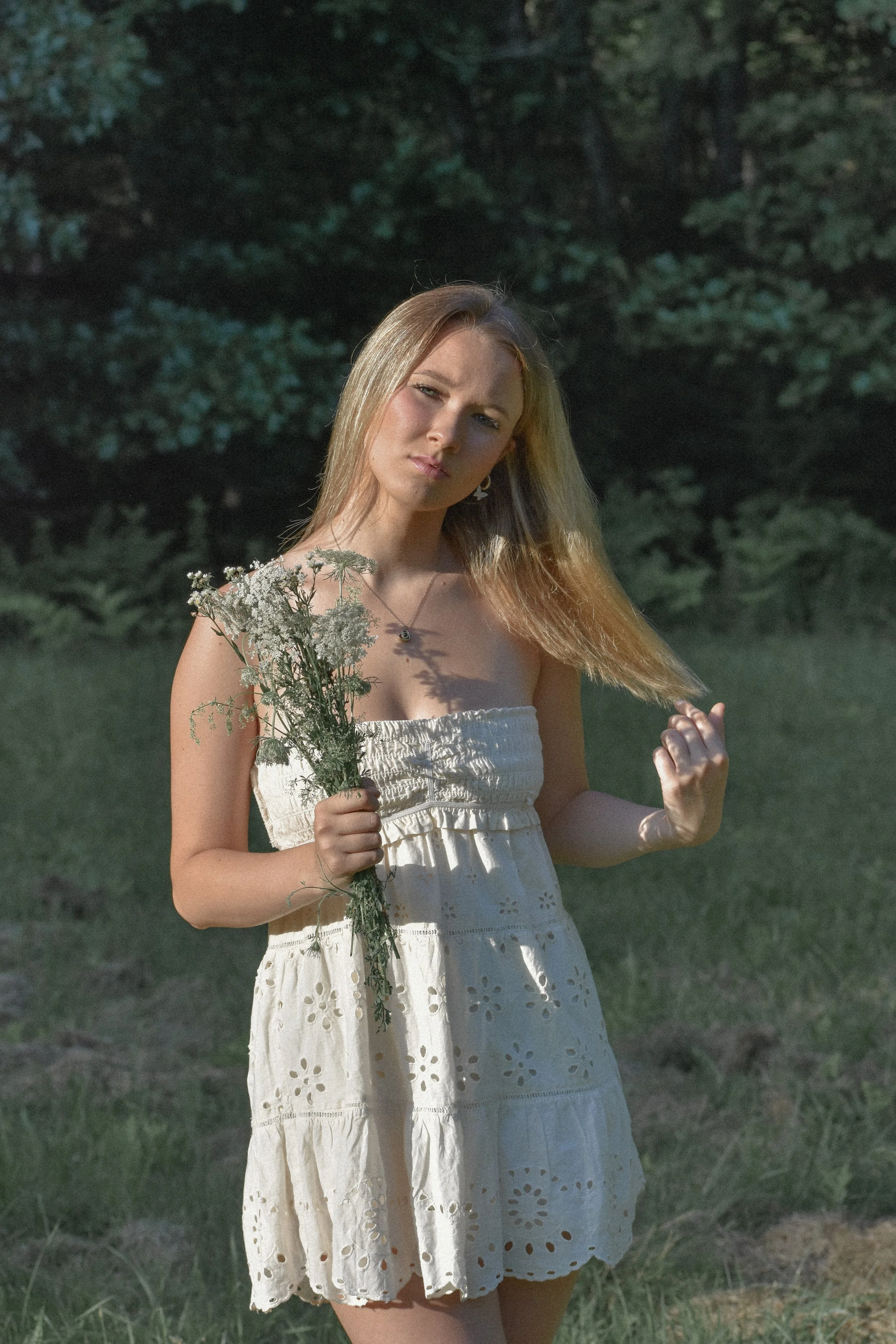 A young woman standing outdoors in a grassy area with trees in the background, wearing a white strapless dress and holding a bouquet of wildflowers.