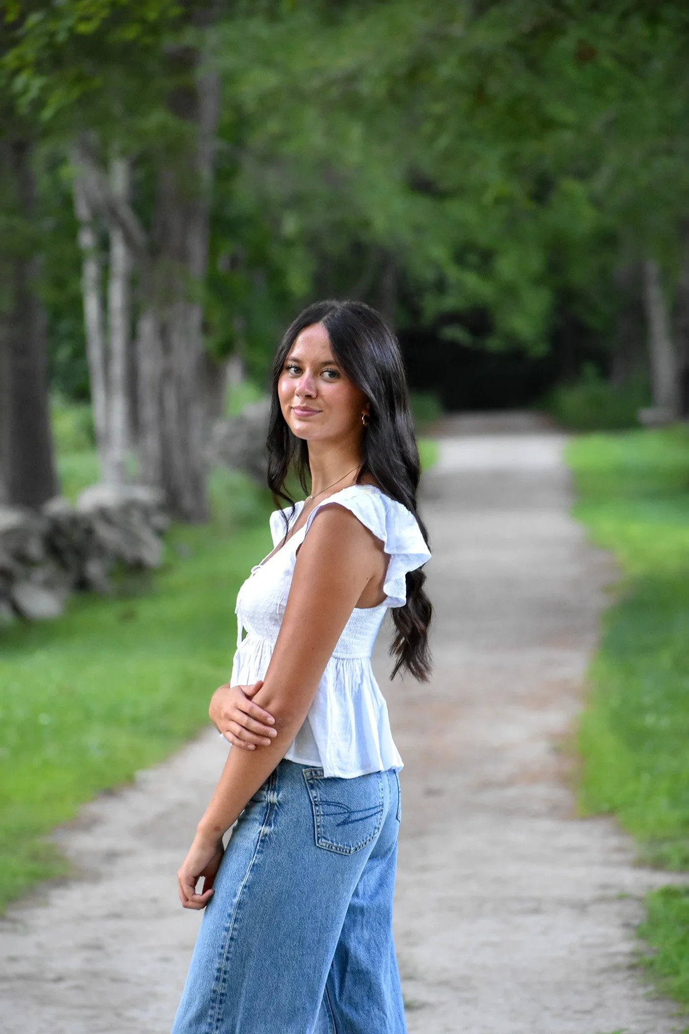 A woman with long dark hair, wearing a white ruffled top and blue jeans, standing on a dirt path in a green, wooded park.