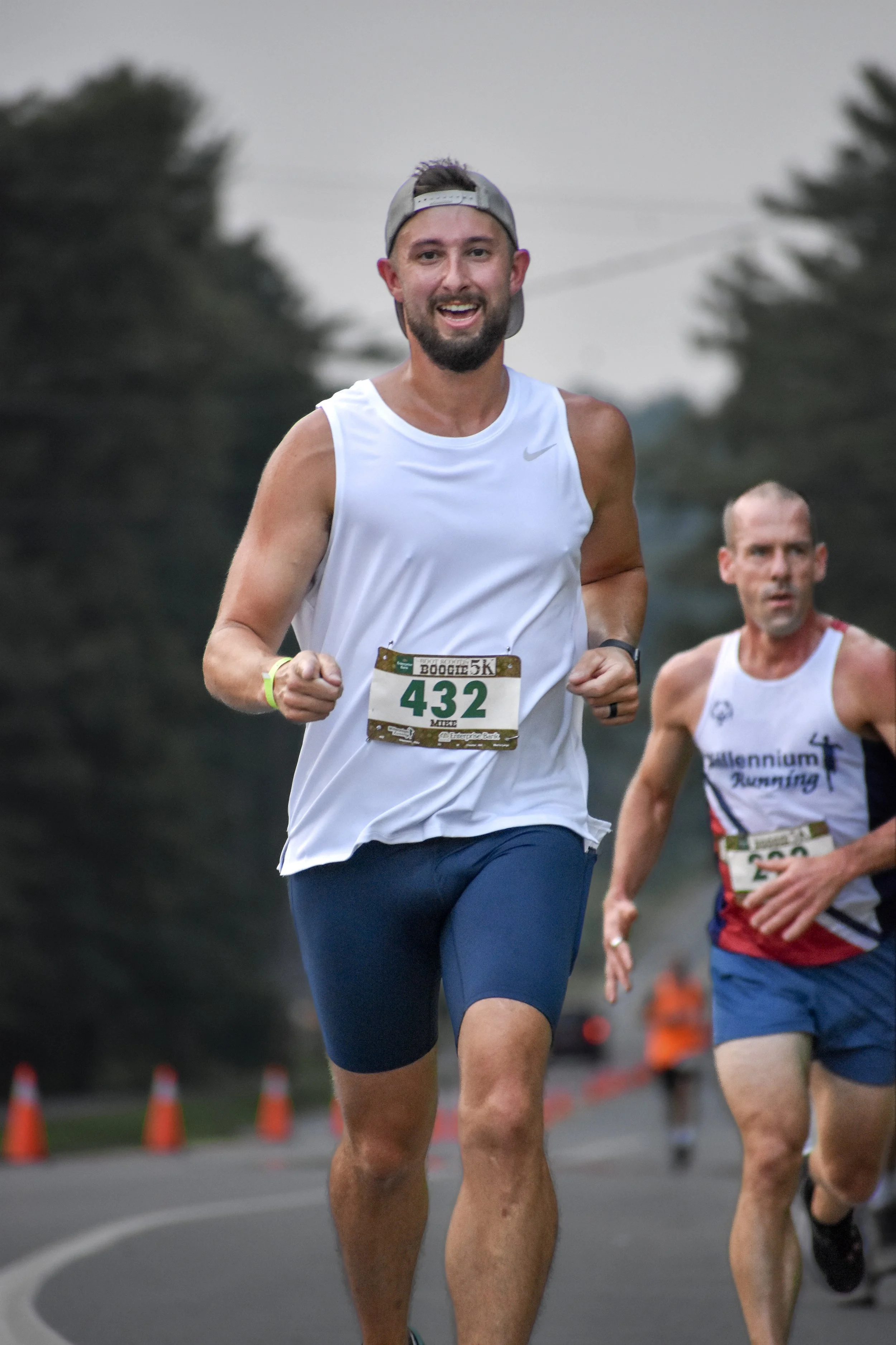 A man running in a road race, wearing a white sleeveless shirt, blue shorts, a gray cap, and a numbered bib 432.