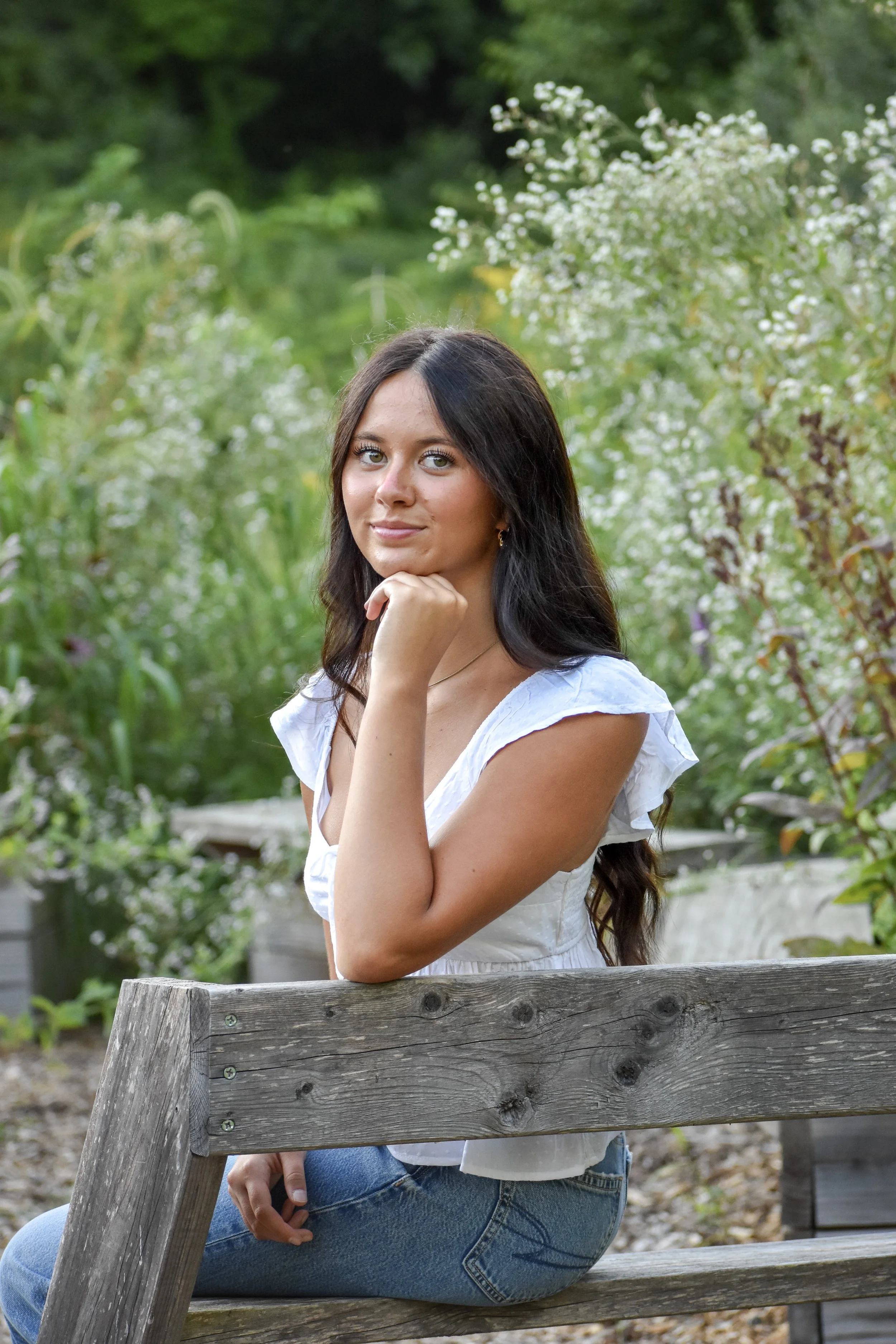 A young woman with long dark hair and blue eyes sits on a wooden bench outdoors, surrounded by greenery and white flowers, resting her chin on her hand and smiling slightly at the camera.