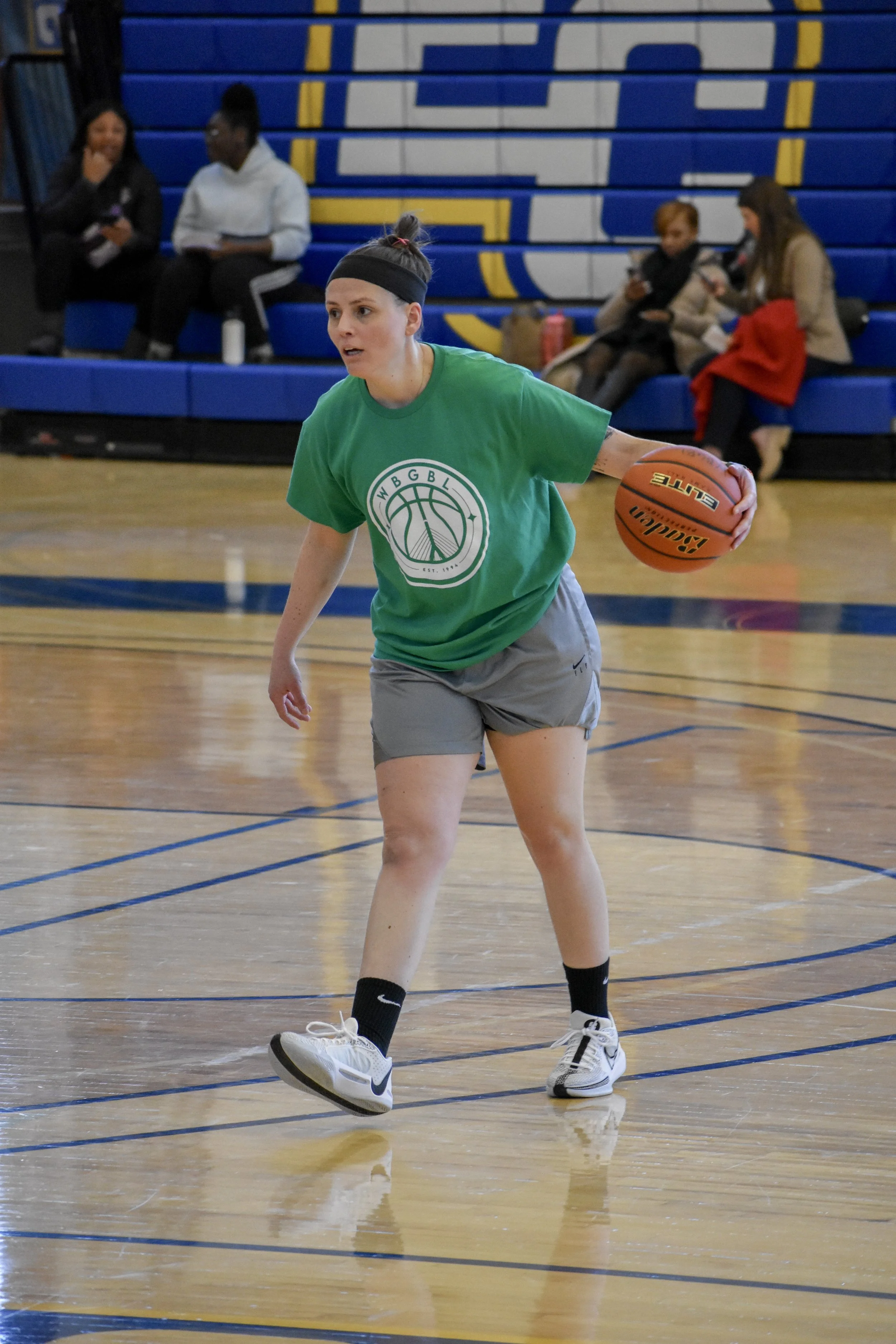 A woman in a green T-shirt and gray shorts playing basketball on an indoor court, with spectators sitting on blue bleachers in the background.