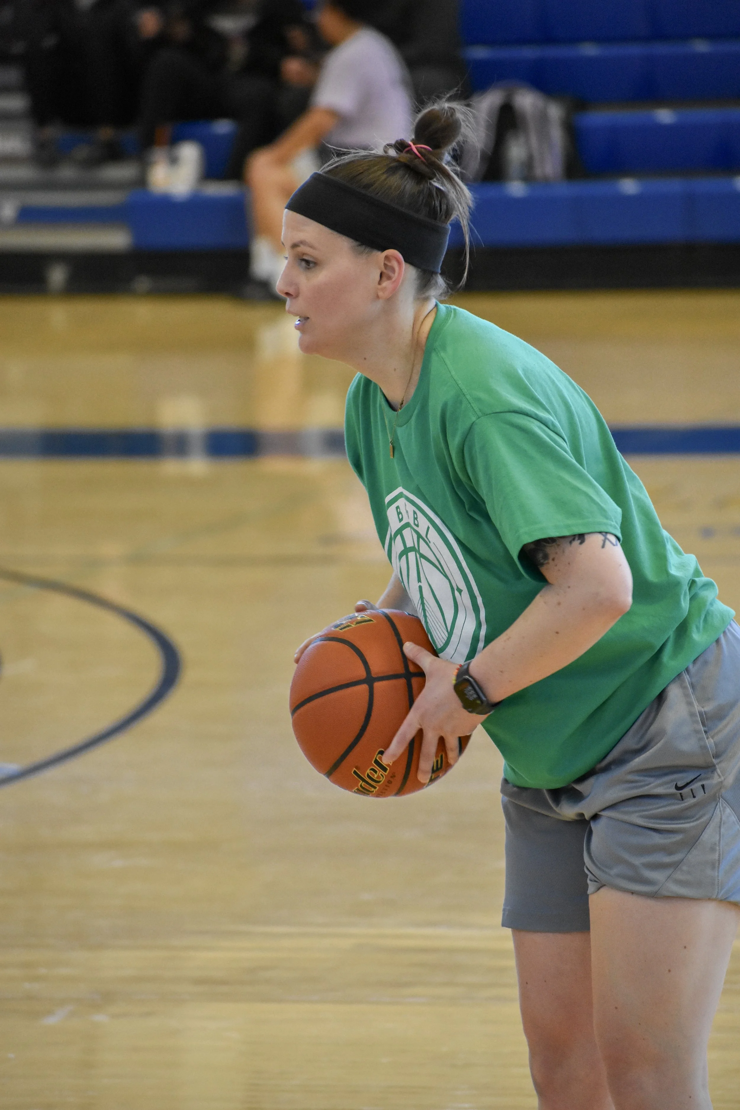 A woman in green shirt and gray shorts holding a basketball on an indoor basketball court.