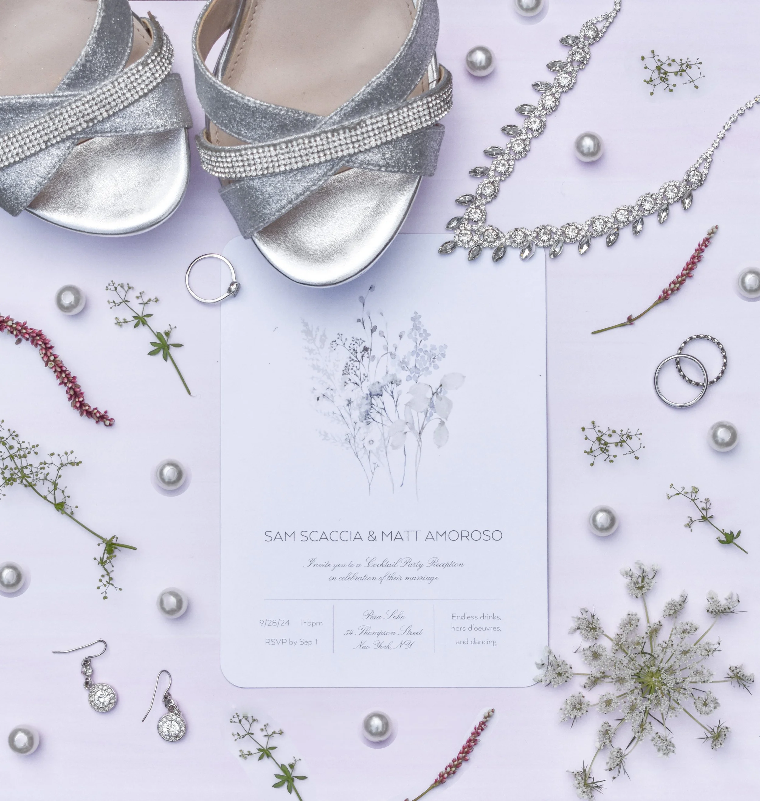 A wedding invitation surrounded by silver high-heeled sandals, pearl jewelry, wedding rings, and small flowers and decorative beads on a light-colored background.