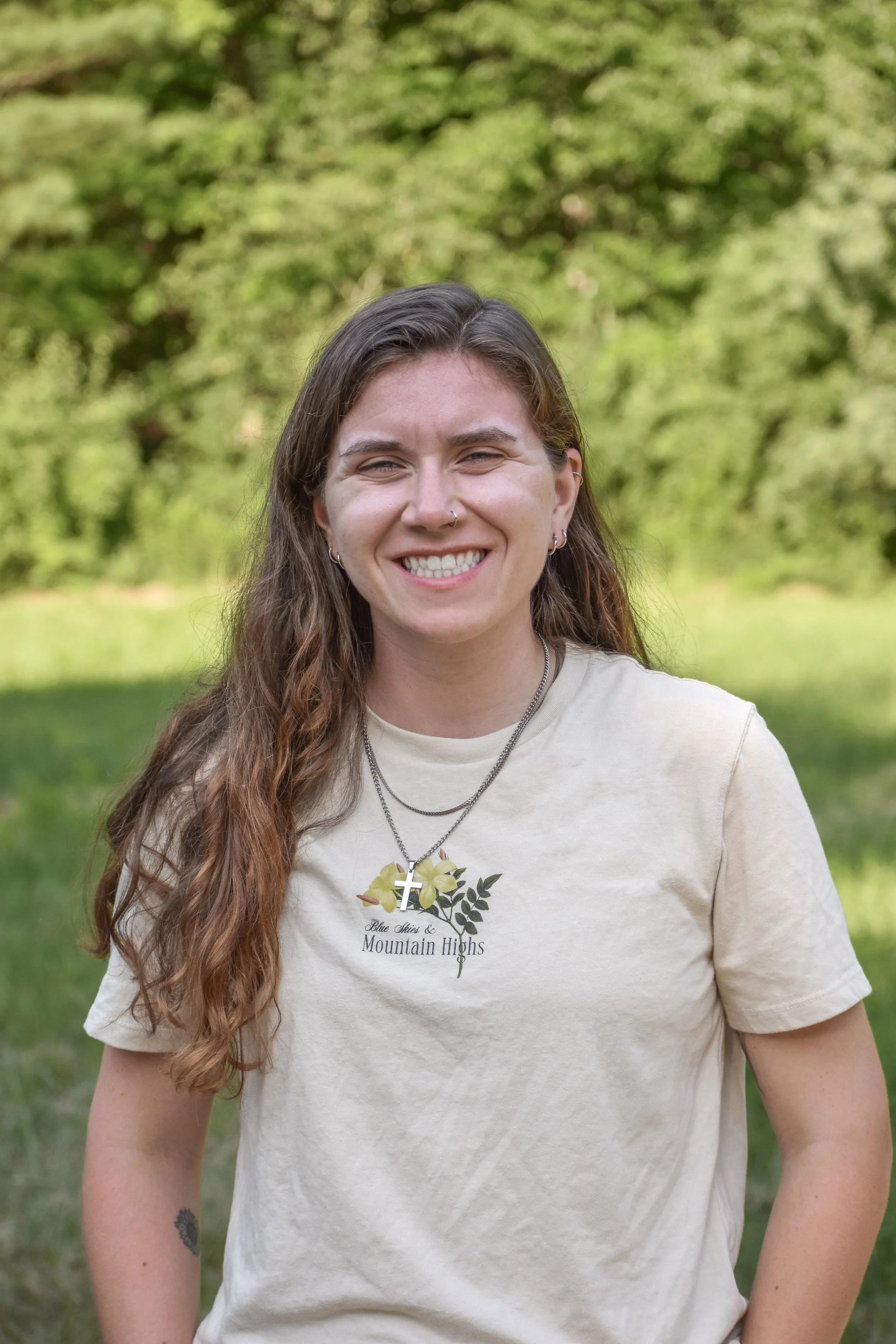A young woman with long, wavy brown hair smiling outdoors, wearing a beige T-shirt and layered necklaces, with a green, leafy background.