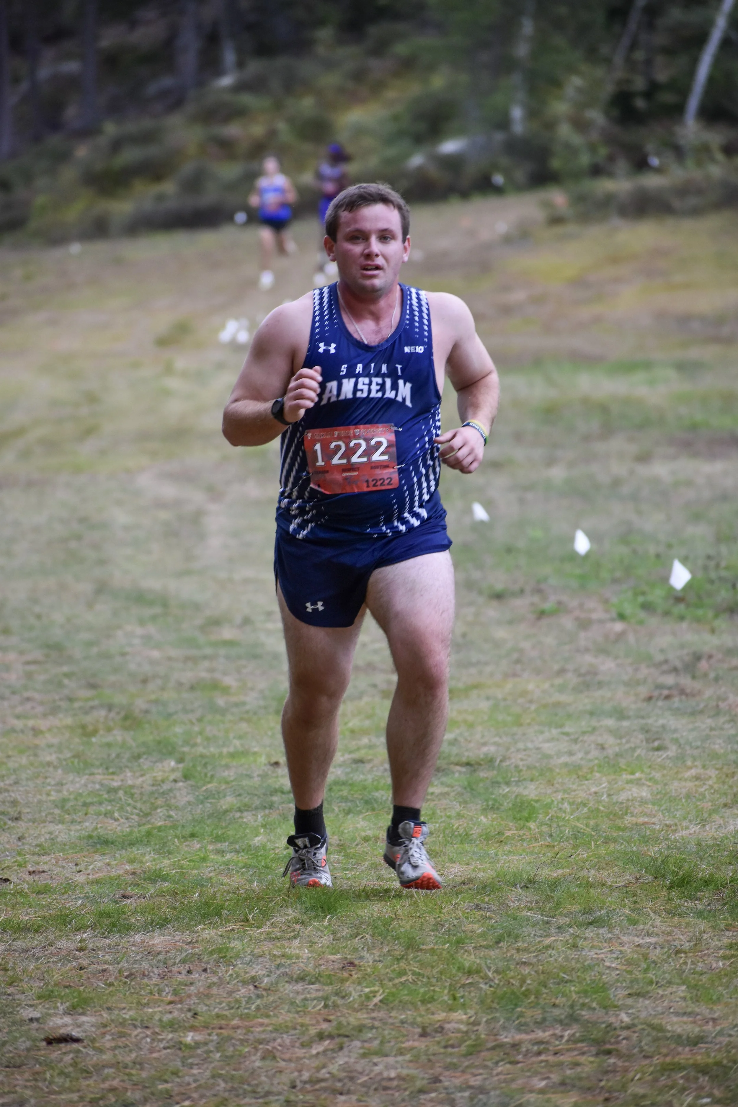 Male runner in blue Saint Anselm race uniform running on grassy trail during a cross-country race with others in the background.