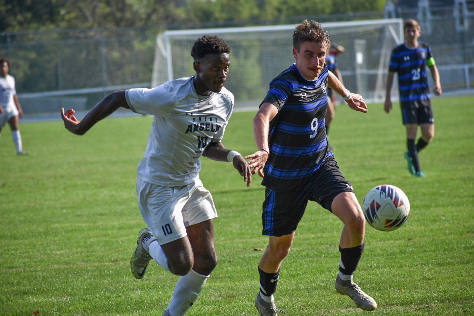 Two soccer players racing for the ball on a grassy field, with other players in the background and a goalpost.