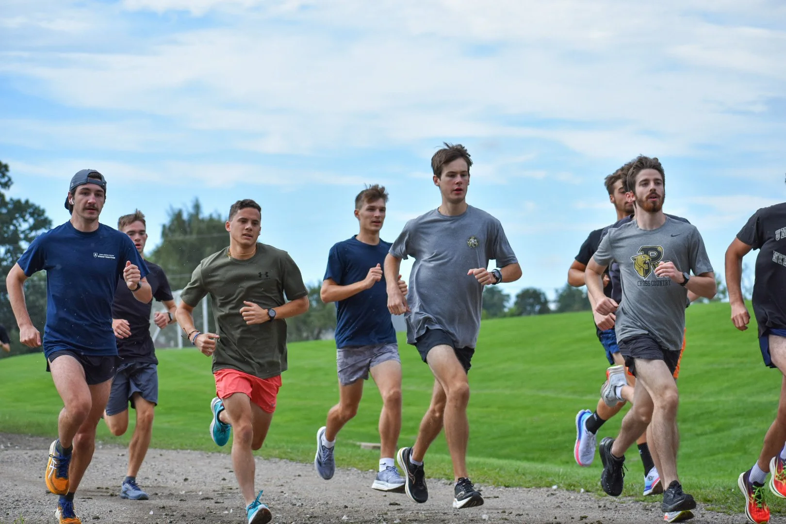 A group of young men running outdoors on a grassy field with a dirt track under a partly cloudy sky.