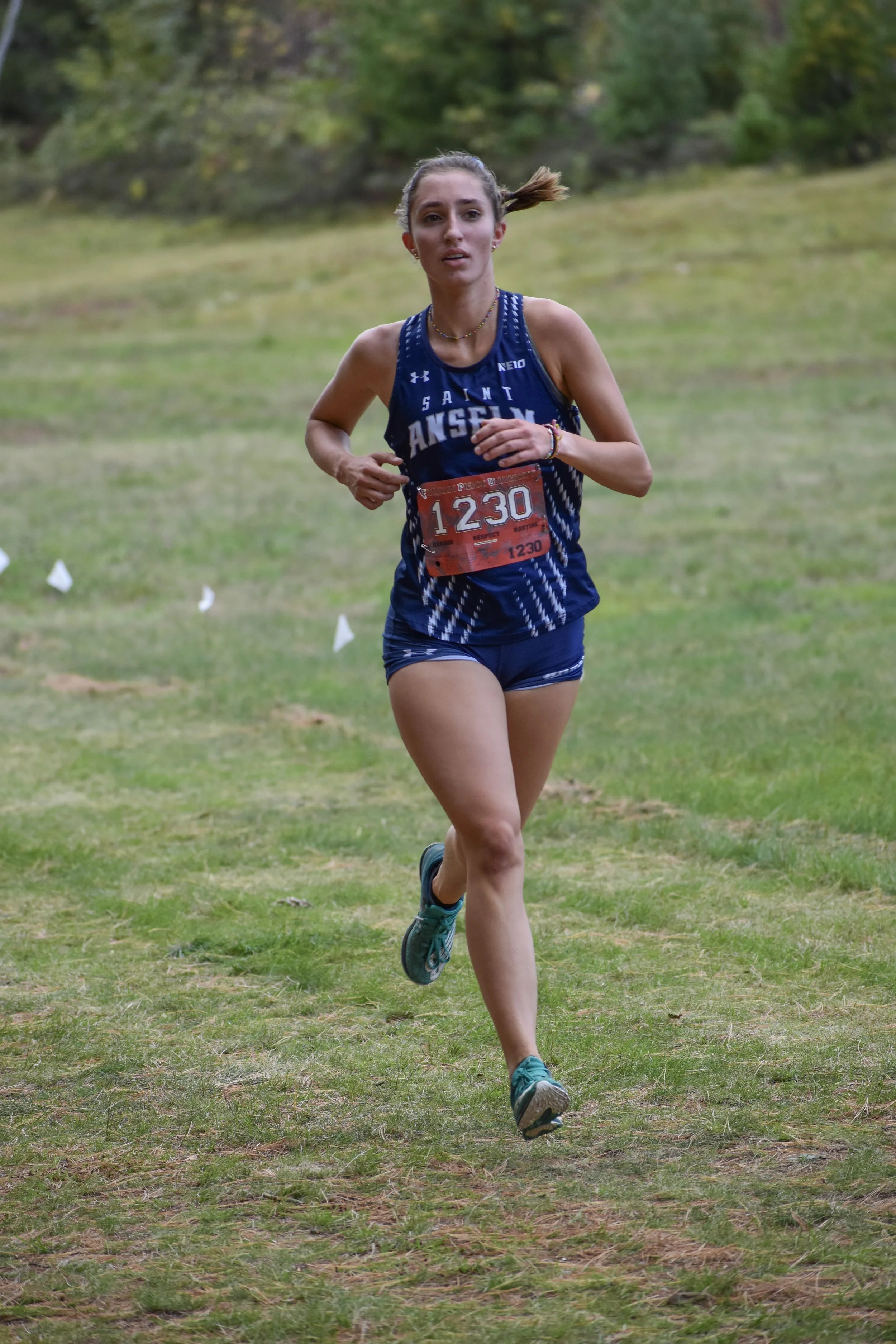 A female runner in a blue athletic uniform with the word 'Saint Anselm' on the front, wearing race bib number 1230, running outdoors on a grassy trail with trees in the background.