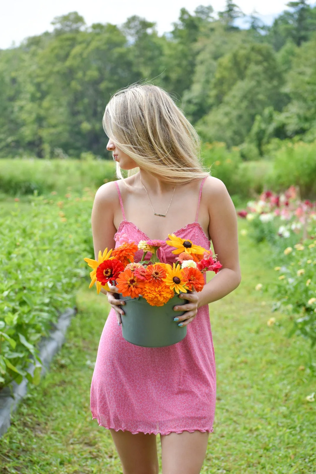 A young woman in a pink dress holding a gray pot of colorful flowers in a lush green garden.
