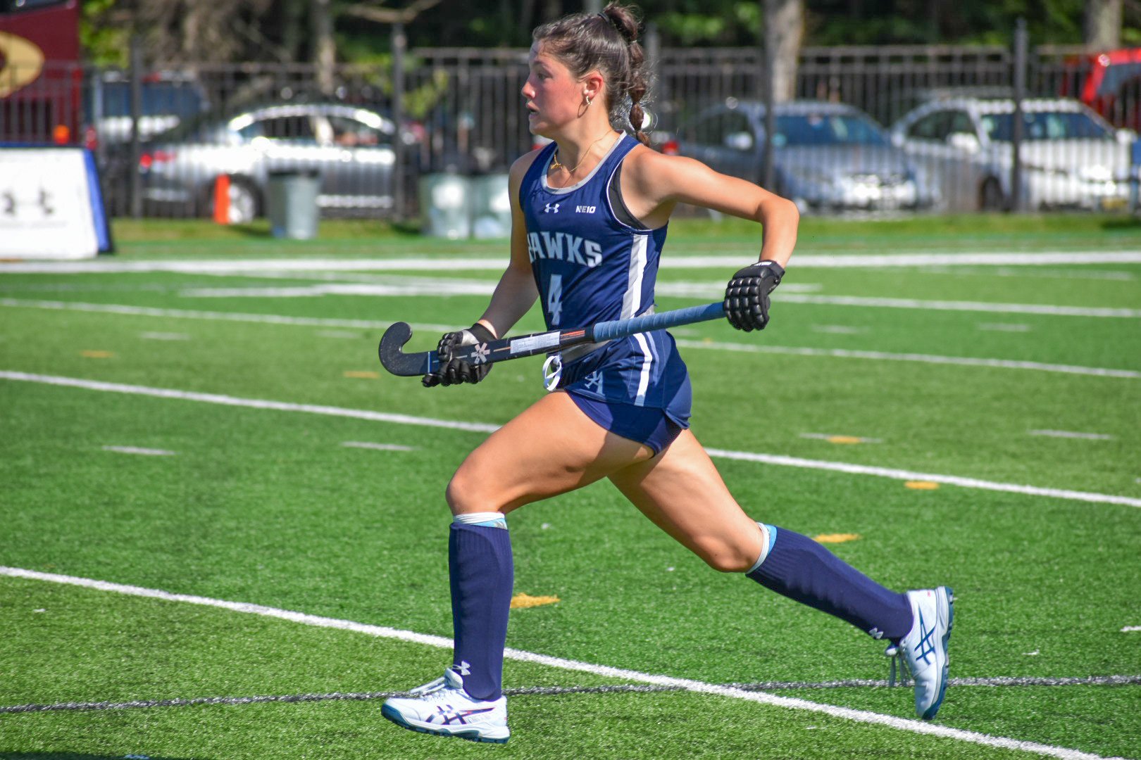A female field hockey player in a blue uniform running on a green turf field holding a field hockey stick.